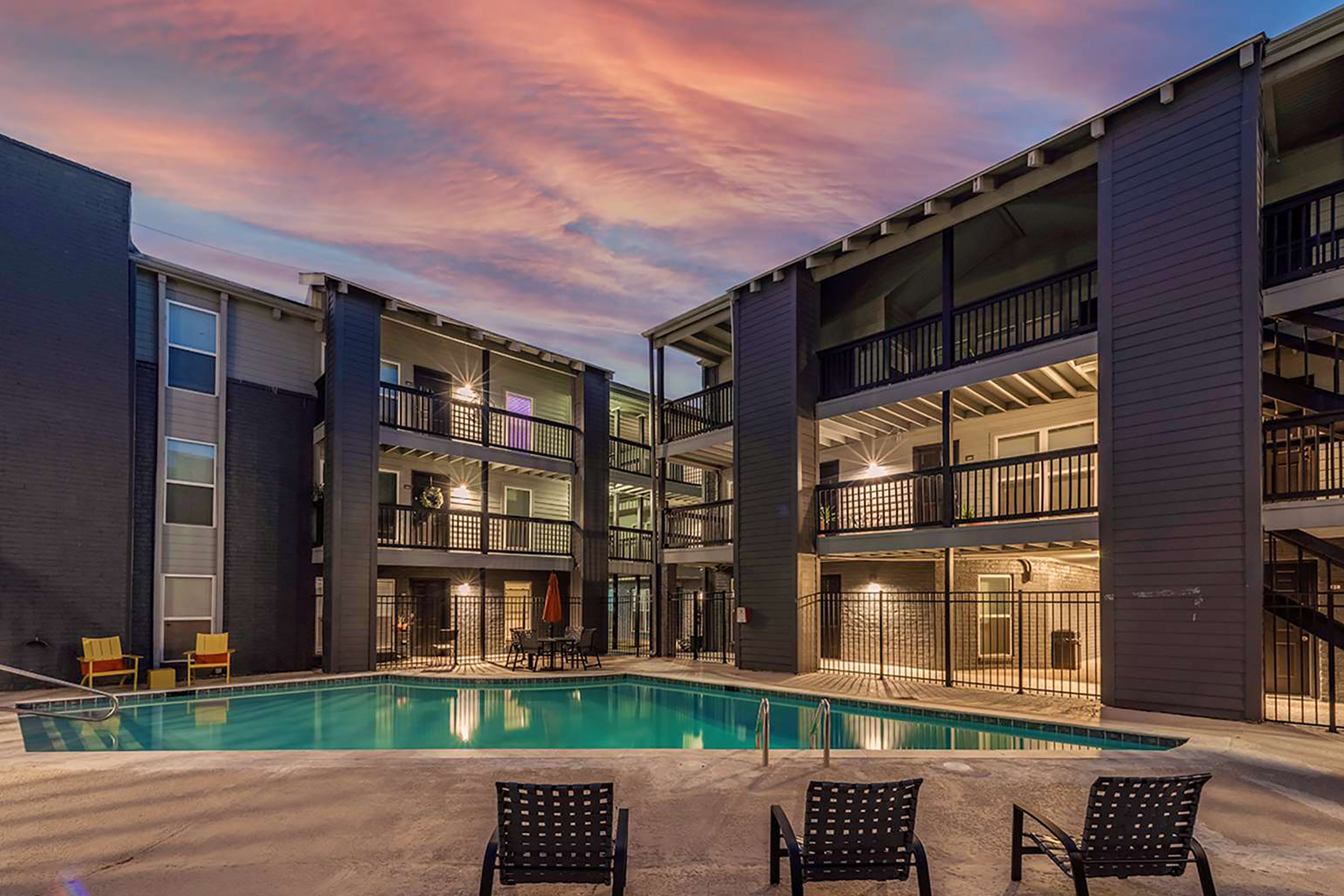 A modern apartment complex featuring two buildings surrounding a swimming pool. The pool area has lounge chairs, and the sky is adorned with vibrant hues of orange and pink during sunset. The buildings have balconies and outdoor furniture, creating a welcoming atmosphere for residents.