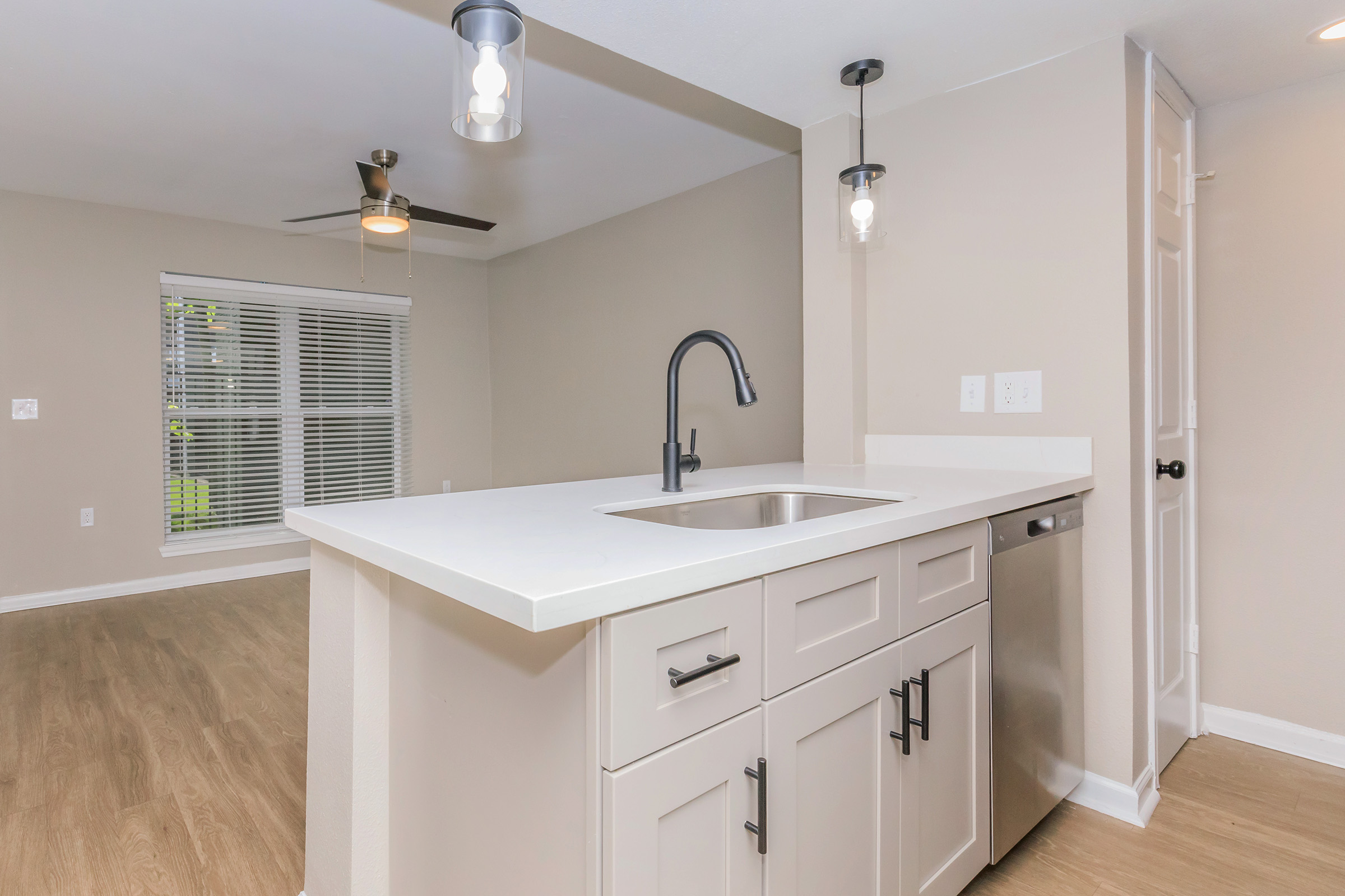 Modern kitchen featuring a sleek countertop and a stainless steel sink. There are decorative pendant lights above the counter, light-colored cabinetry, and a doorway leading to a spacious living area with large windows. The flooring is a warm, wood-like laminate.