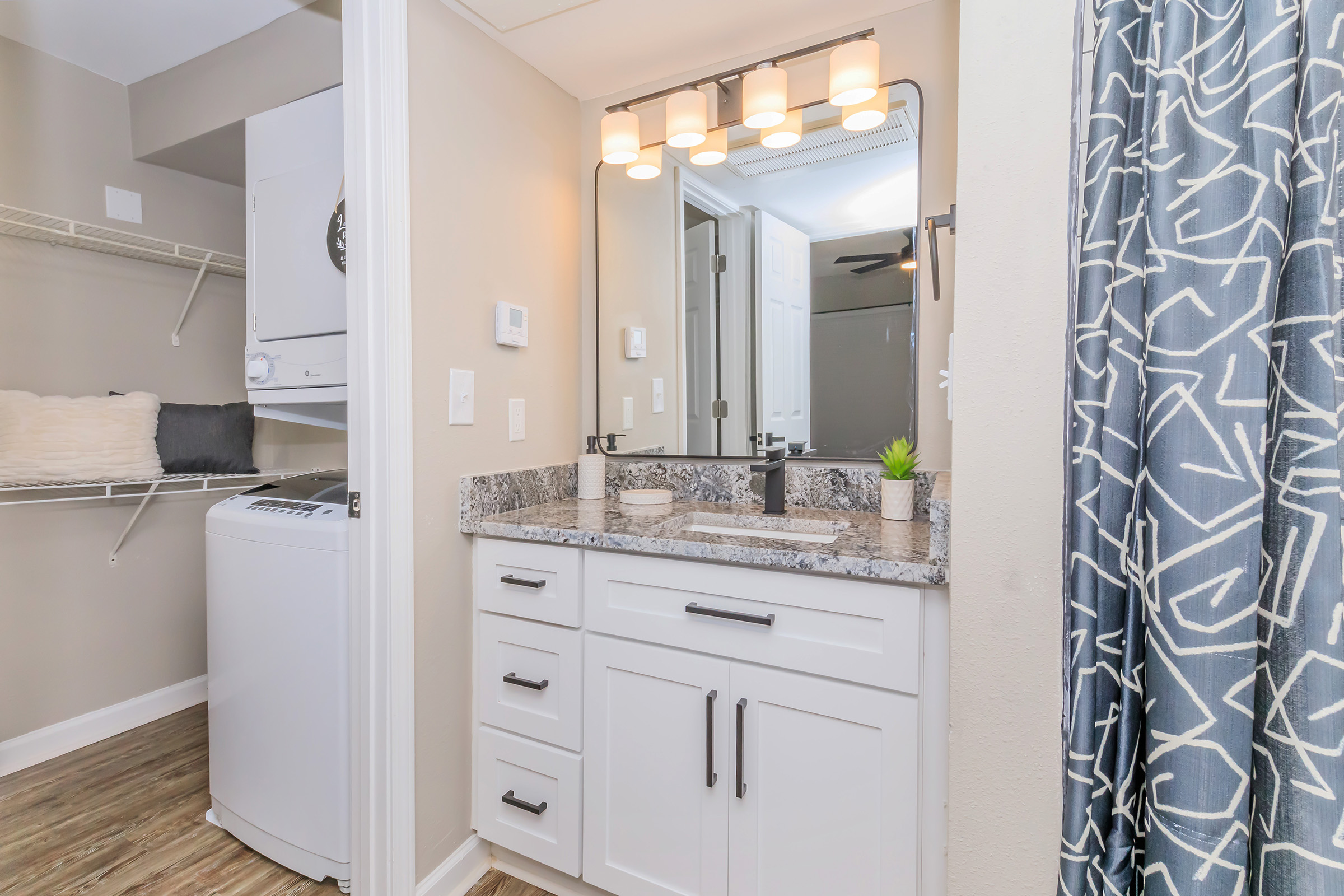 A modern bathroom with a stylish vanity featuring a gray granite countertop, black handles, and a large mirror above. There is a light fixture with multiple bulbs. To the side, a laundry area is visible with a washing machine and shelving. The decor includes a plant and a patterned curtain.