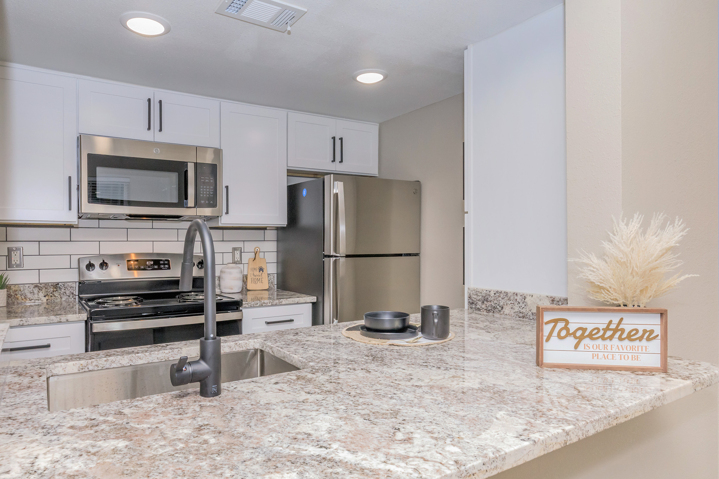 A modern kitchen featuring white cabinetry, stainless steel appliances, and a granite countertop. A sink with a sleek faucet is in the foreground, while the background includes a stove, microwave, and refrigerator. A decorative sign reading "Together is our favorite place to be" is on the countertop, alongside a small plant and dishware.
