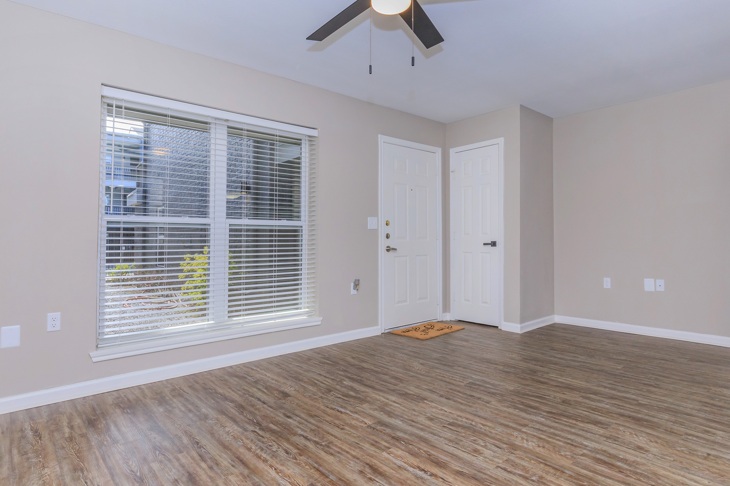 A spacious, empty room featuring light-colored walls and a wooden floor. There is a large window with blinds allowing natural light, and a ceiling fan is mounted above. The room has a doorway leading to another area, with a welcome mat at the entrance.