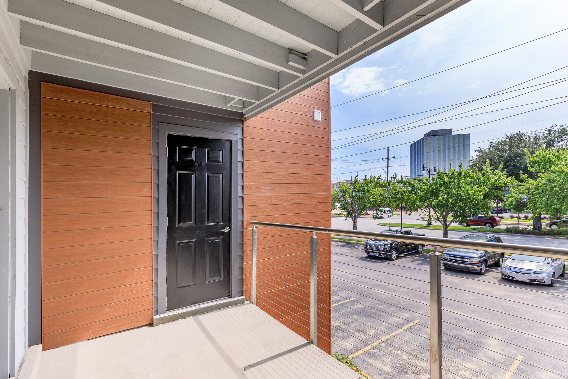 A balcony view featuring a black door against a wooden wall, with a railing in the foreground. In the background, there's a modern building and a parking lot filled with cars under a clear blue sky with some clouds. Trees line the street, adding greenery to the scene.