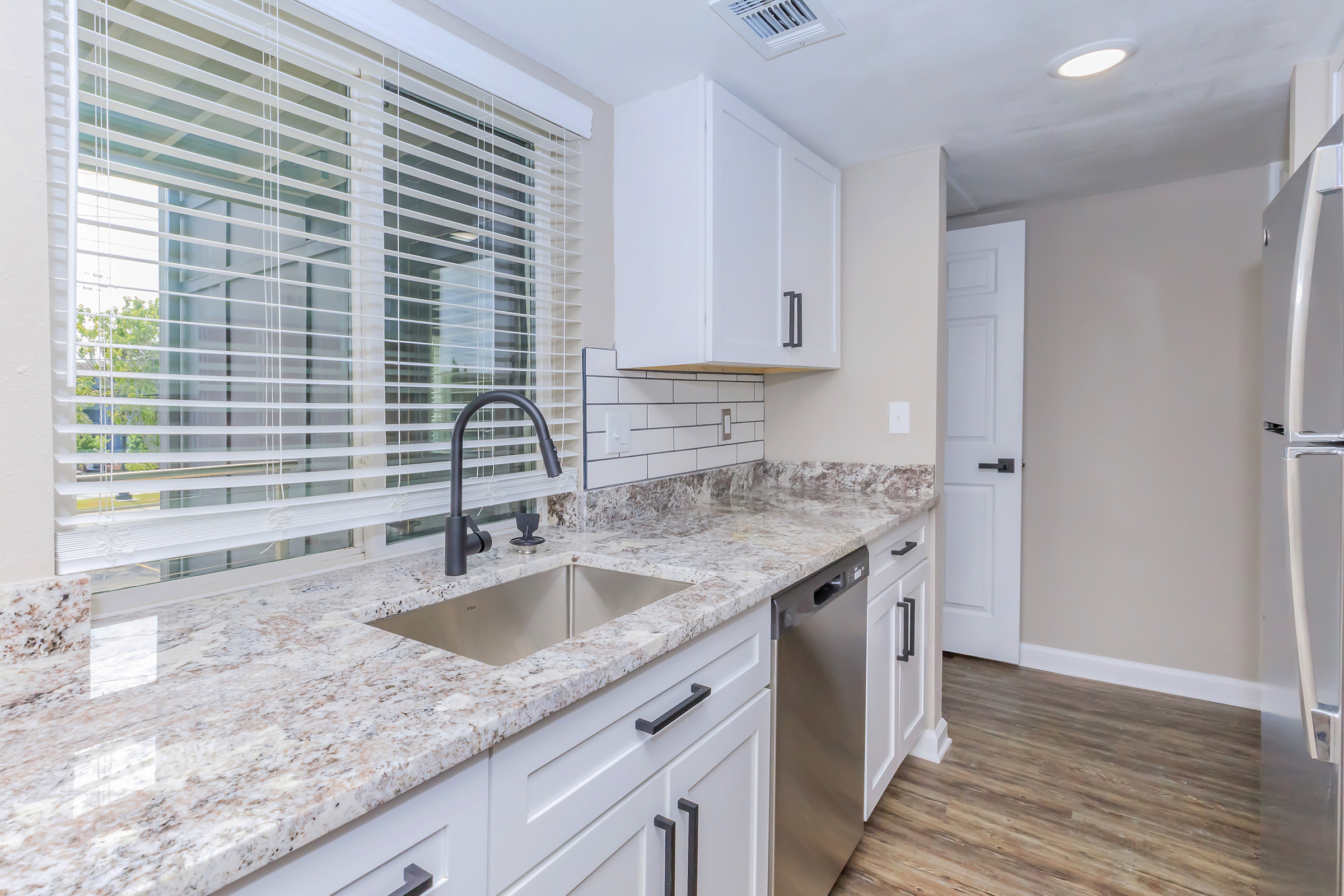 Modern kitchen featuring white cabinetry, a stainless steel dishwasher, a granite countertop with an undermount sink, and a tile backsplash. Large window with blinds allows natural light. Floor is a warm, wood-like laminate. Open layout with a doorway leading to an adjacent room.