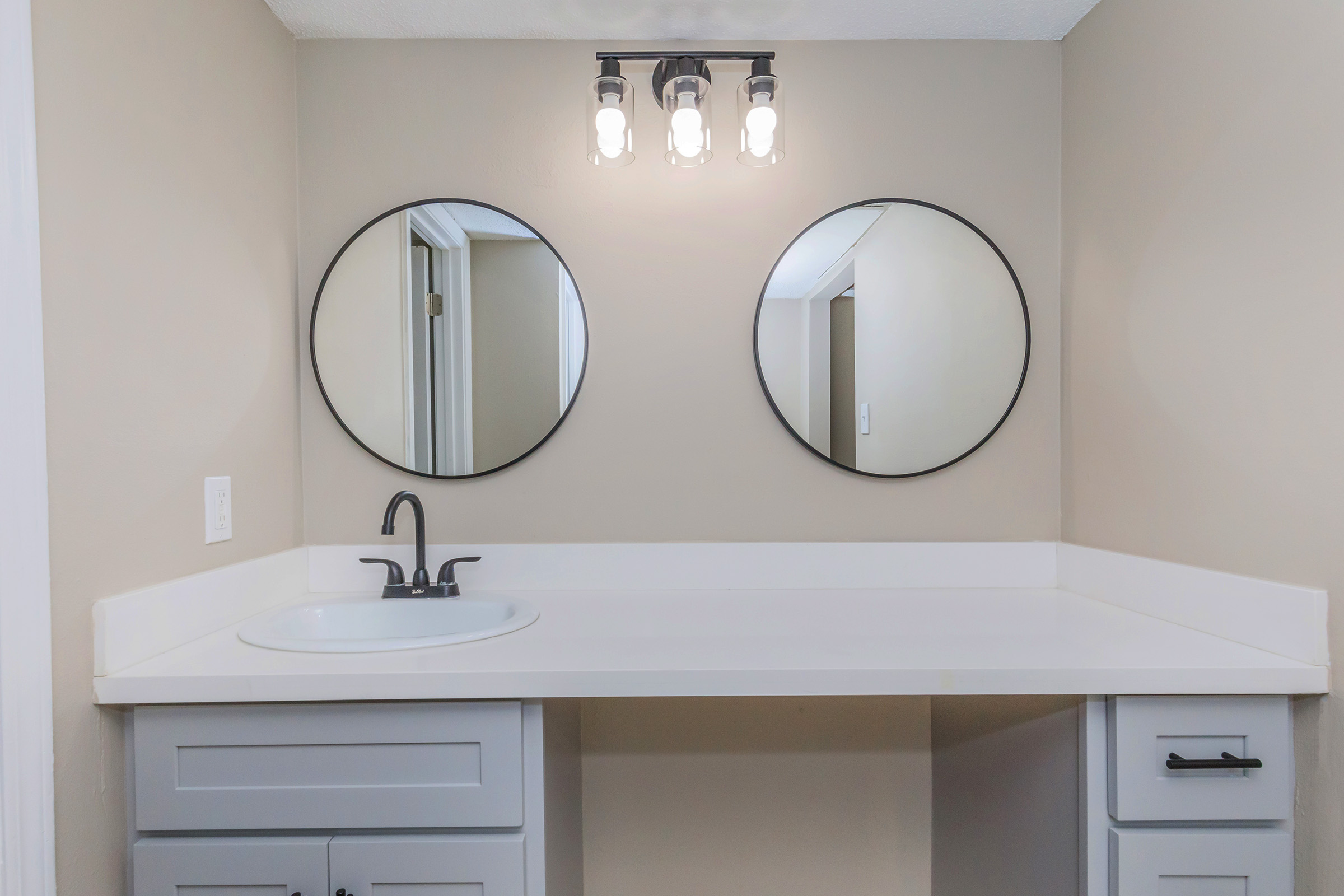 A modern bathroom vanity featuring a white countertop with a single sink, two round mirrors above the sink, and a stylish light fixture with three bulbs. The walls are painted a soft beige, and there are grey cabinets underneath the countertop. The overall design is clean and contemporary.
