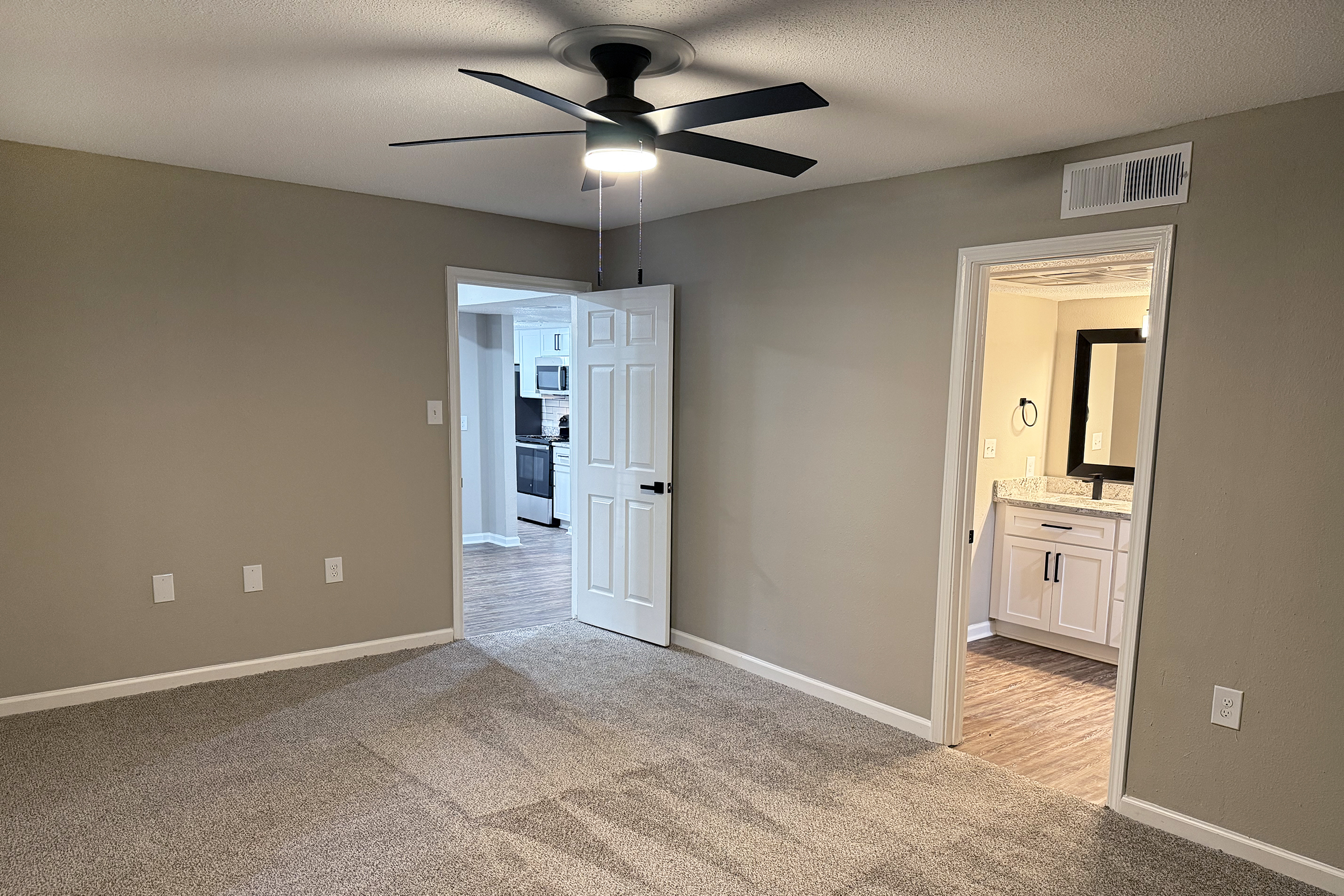 A spacious room with light brown carpet, a ceiling fan, and two doorways leading to a bathroom and a kitchen. The walls are painted in a warm beige color. Natural light is provided from the nearby openings, enhancing the cozy atmosphere of the space.