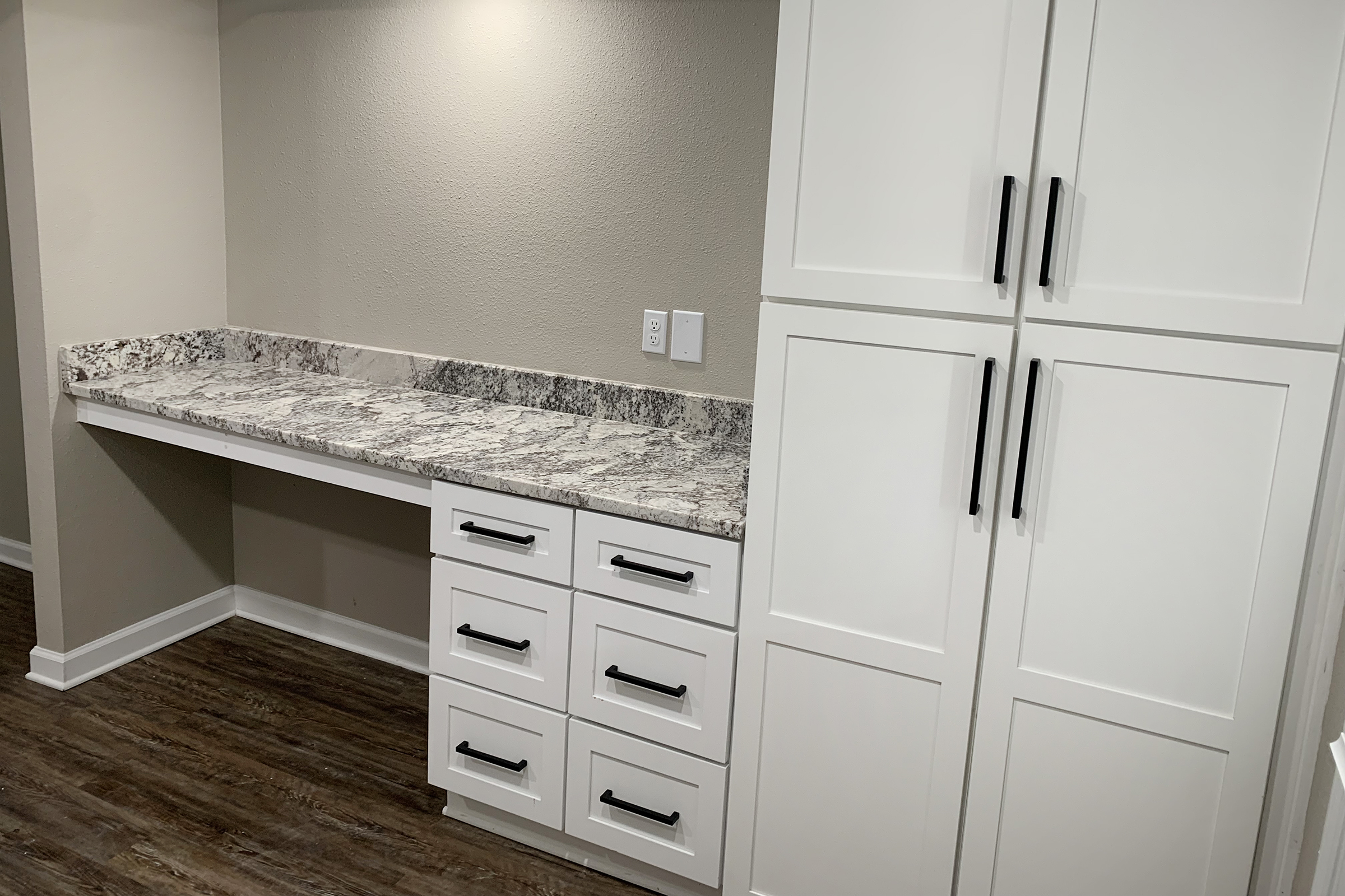 A modern kitchen workspace featuring a sleek granite countertop with a subtle pattern, situated next to white cabinetry. The cabinetry includes multiple drawers with black handles, and adjacent is a tall cabinet with matching handles, all set against a neutral wall and wooden floor.