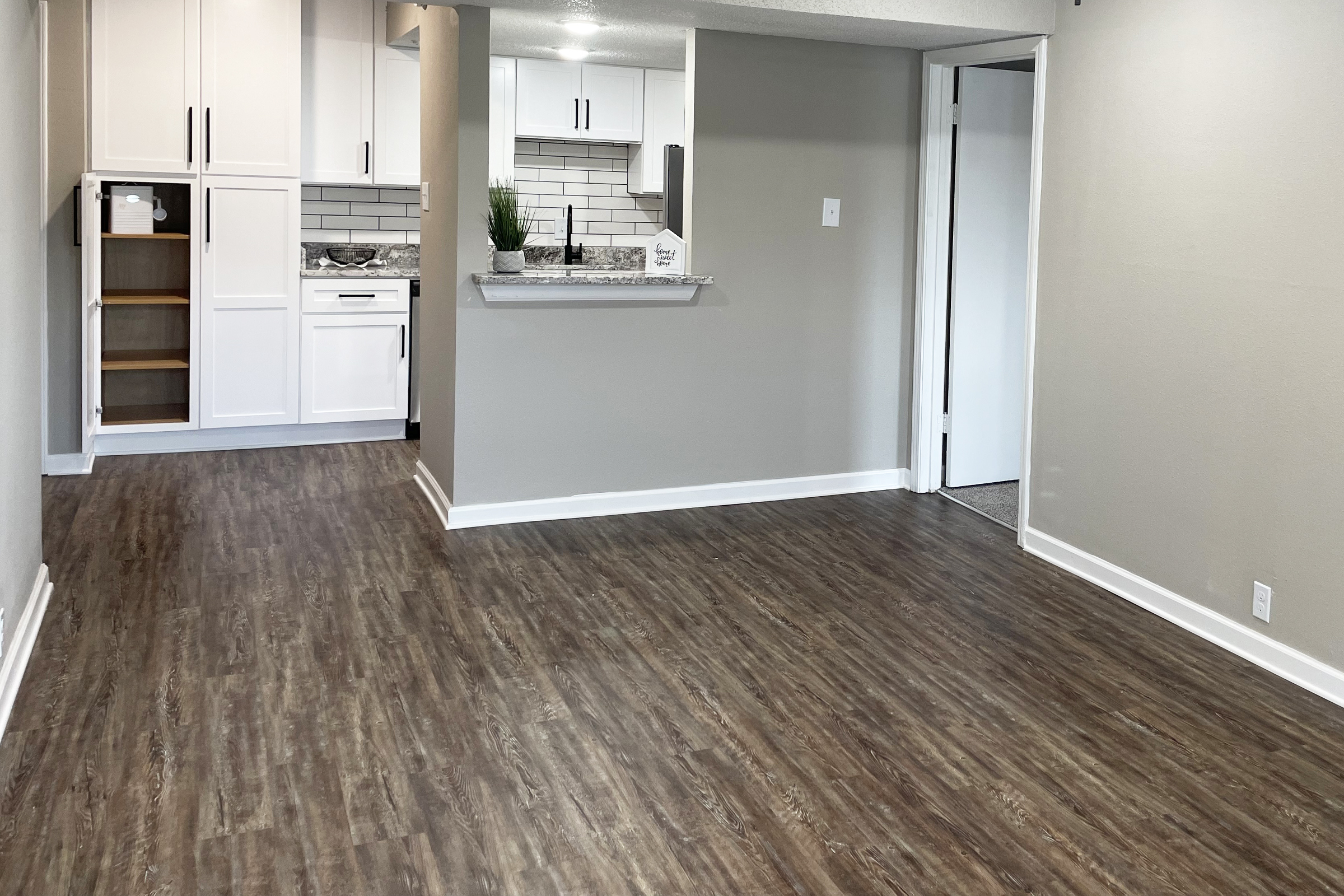 Interior of a modern apartment featuring an open layout. The living area has wood-like flooring and fresh gray walls. A kitchenette with white cabinets and a bar counter is visible in the background. A door leads to another room on the right, and there's a small storage area to the left.