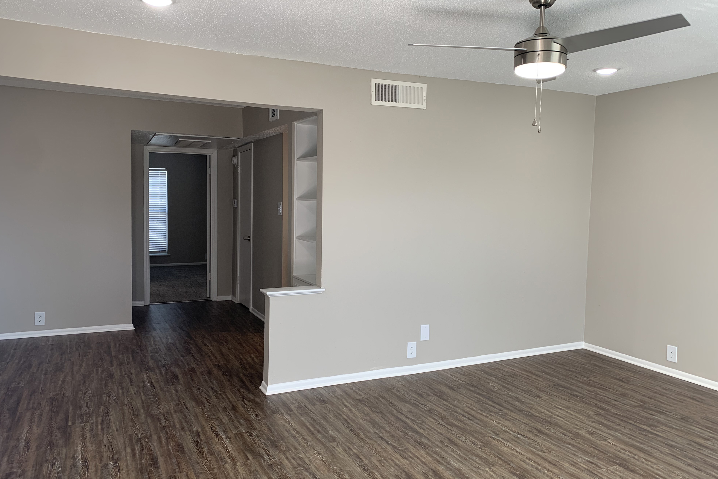 A spacious, lightly colored living room featuring a ceiling fan, hardwood-style flooring, and a small step leading to a hallway. The hallway leads to another room with a window providing natural light. The walls are painted in a neutral tone, creating a modern and inviting atmosphere.
