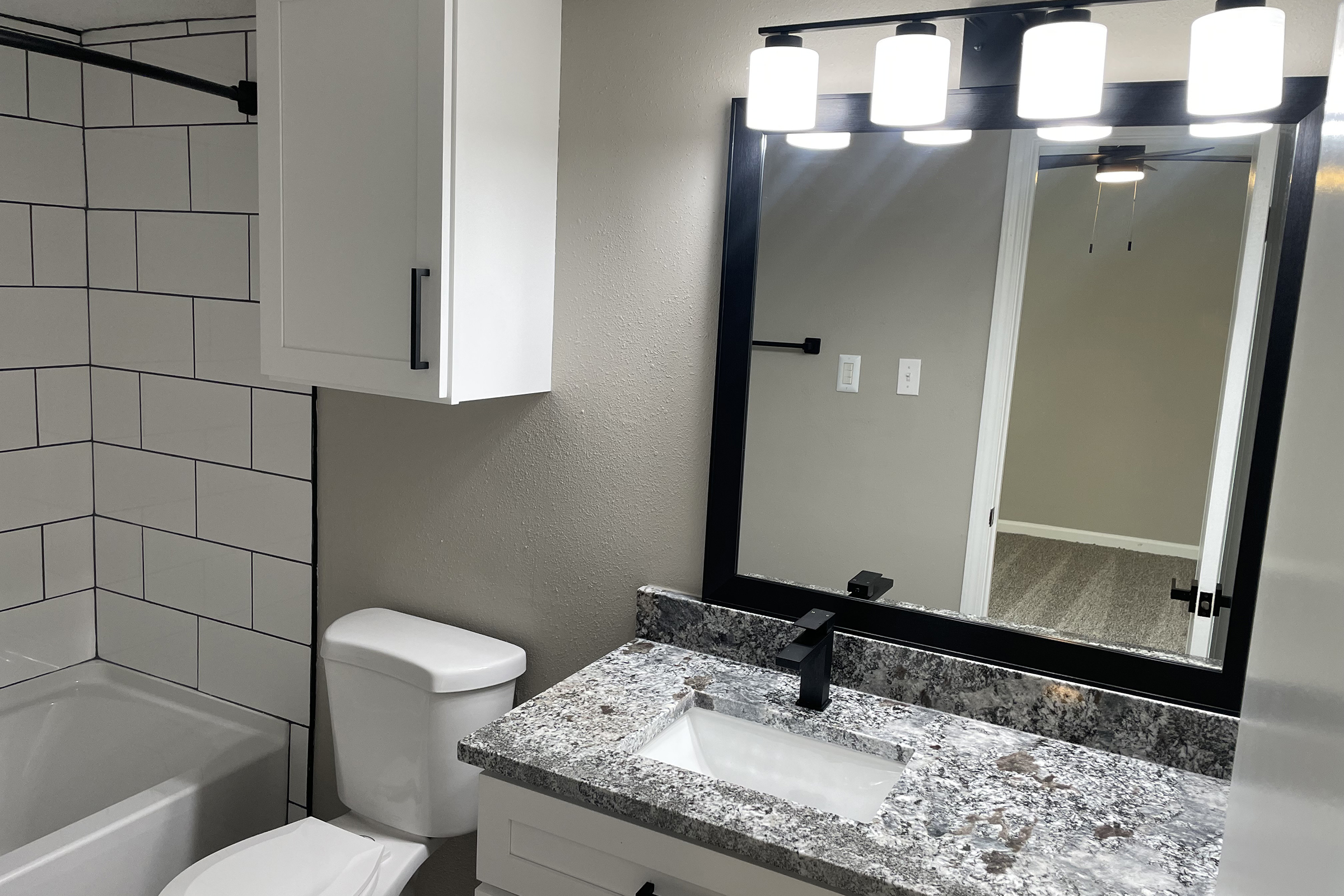 A modern bathroom featuring a white tiled shower, a sleek black-framed mirror above a stone countertop with a rectangular sink. The space includes a white toilet and a light fixture with multiple round bulbs, complemented by neutral wall colors and a doorway leading to another room.