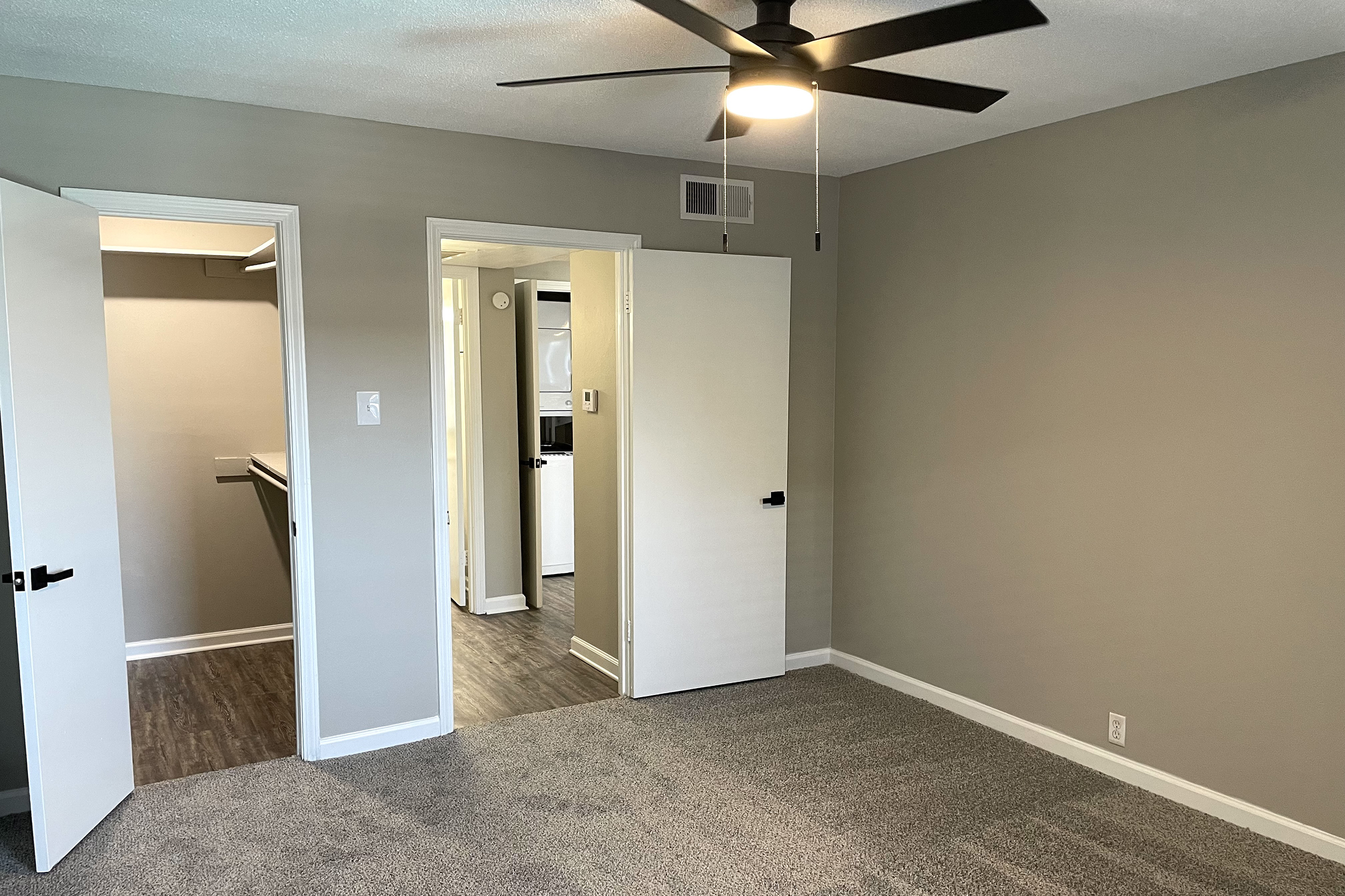 A clean, well-lit room with light grey walls and carpet. There are two open doors: one leading to a closet and another to an adjoining area, possibly a kitchen. A ceiling fan is visible, and natural light enters the space, creating an inviting atmosphere.