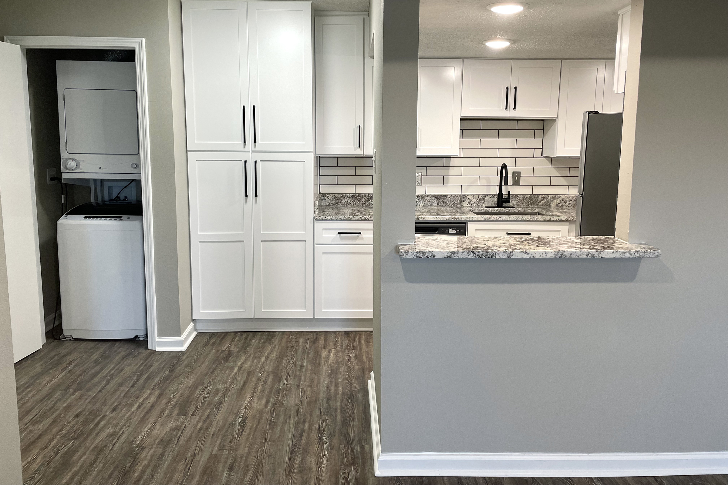 Modern kitchen and laundry area viewed from an adjacent room. The kitchen features white cabinets, a gray and white granite countertop, and a subway tile backsplash. There is a black sink and appliances, including a refrigerator. A laundry unit is visible through an open door. The flooring is a wood-like laminate.