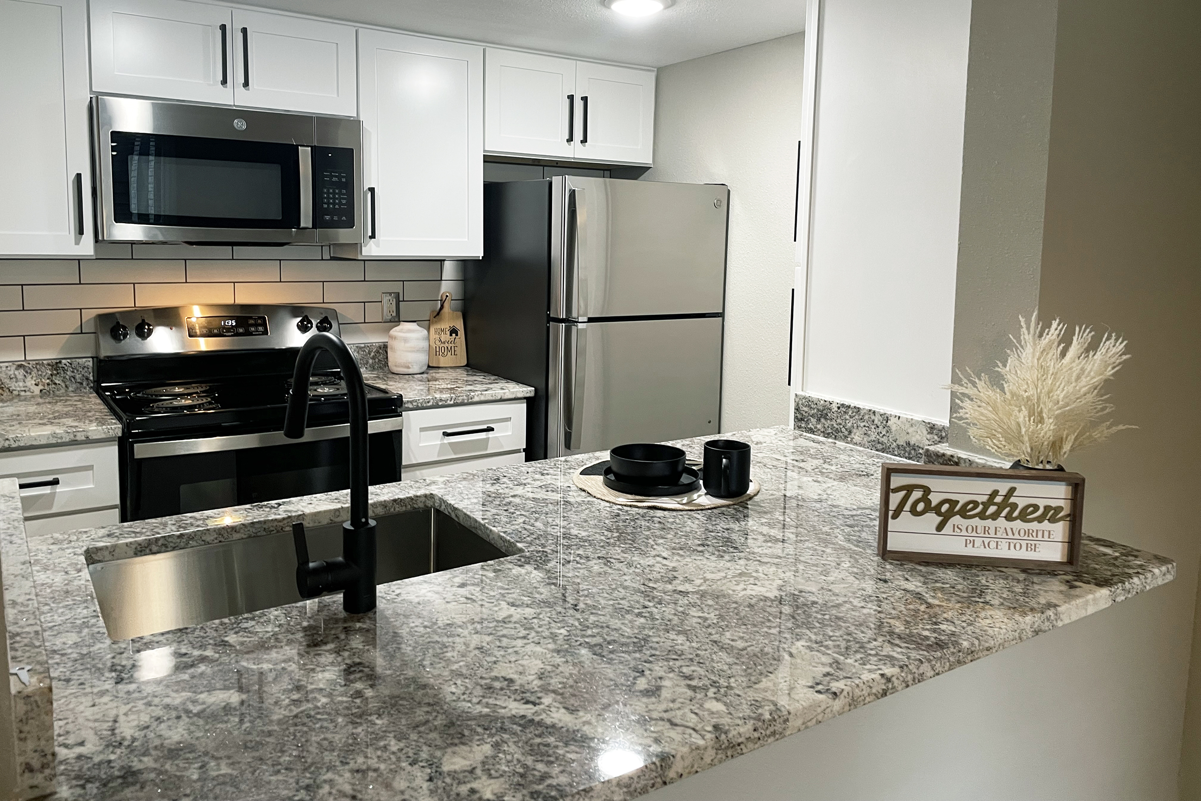 Modern kitchen featuring granite countertops, white cabinetry, stainless steel appliances, and a black sink. A decorative sign reading "Together" sits beside a small vase with dried plants and a black cup on a neutral-colored plate, creating a cozy and inviting atmosphere.
