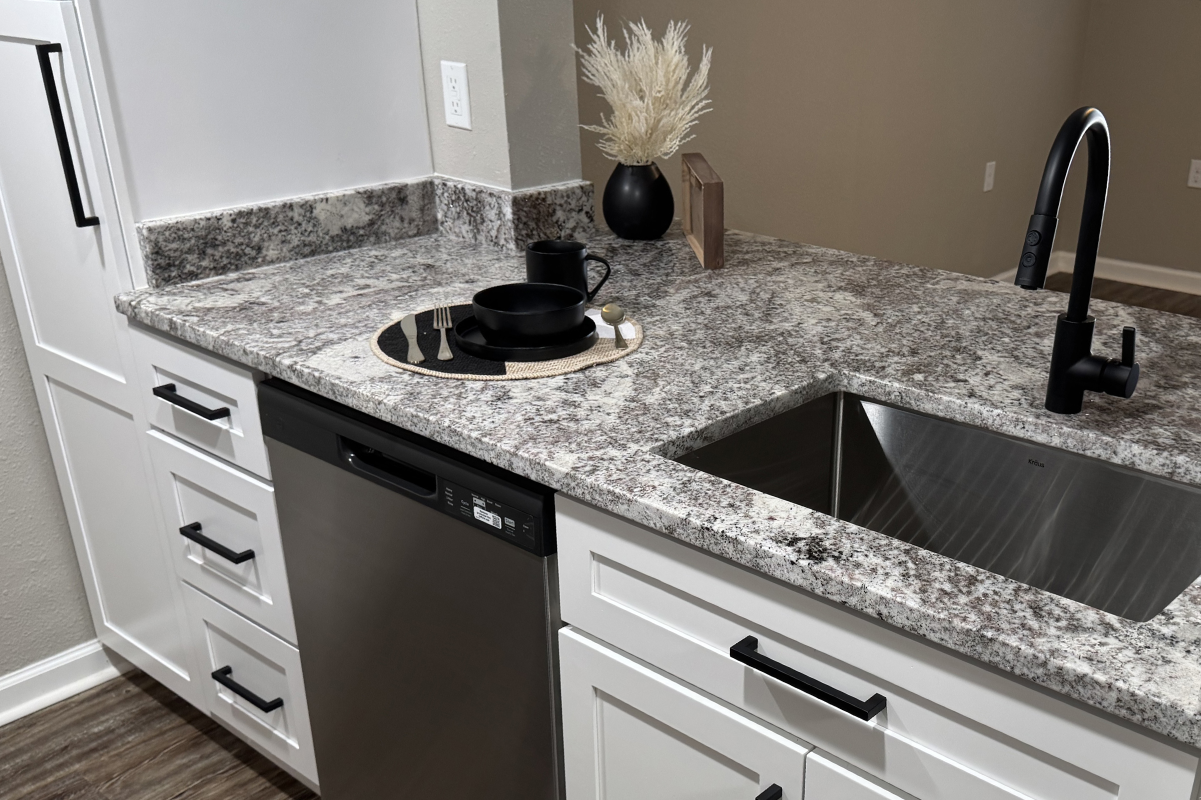 A modern kitchen countertop featuring granite with a stainless steel dishwasher, a sleek black faucet, and minimalist decor. A black mug sits on a round placemat next to a small plate, while a decorative item and dried plant add a touch of elegance to the space.