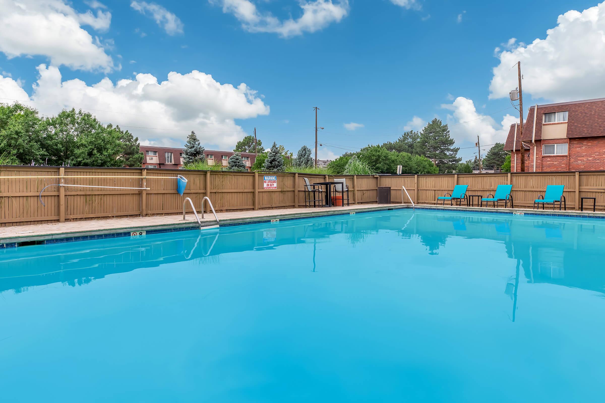 A clear blue swimming pool surrounded by a wooden fence. In the foreground, there are pool ladders and lounge chairs positioned along the deck. The sky is bright with white clouds, and there are bushes and buildings visible in the background.