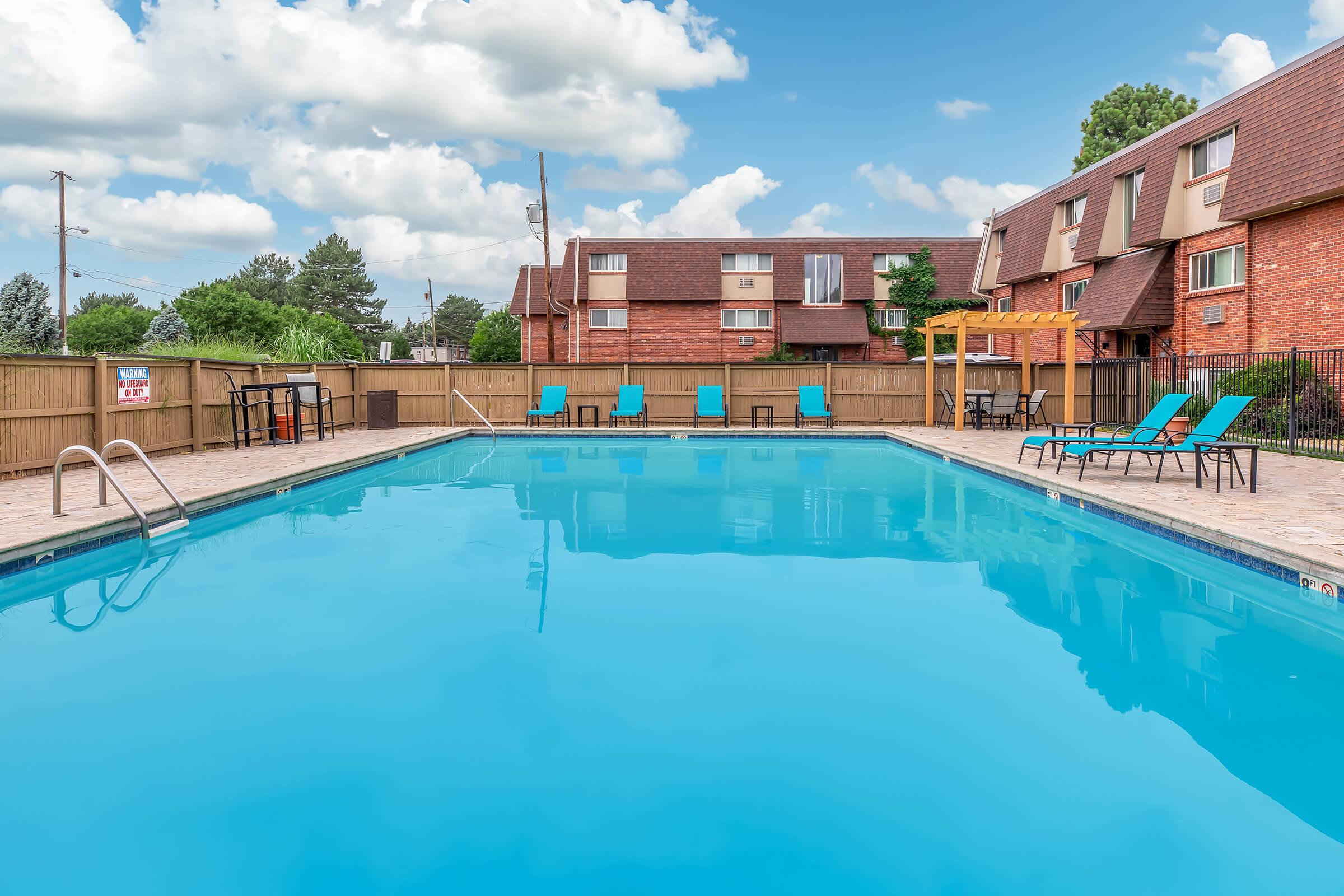 A tranquil outdoor swimming pool surrounded by lounge chairs, with a clear blue sky and fluffy white clouds overhead. The pool area features a wooden pergola and is enclosed by a fence, with nearby brick buildings providing a cozy backdrop.