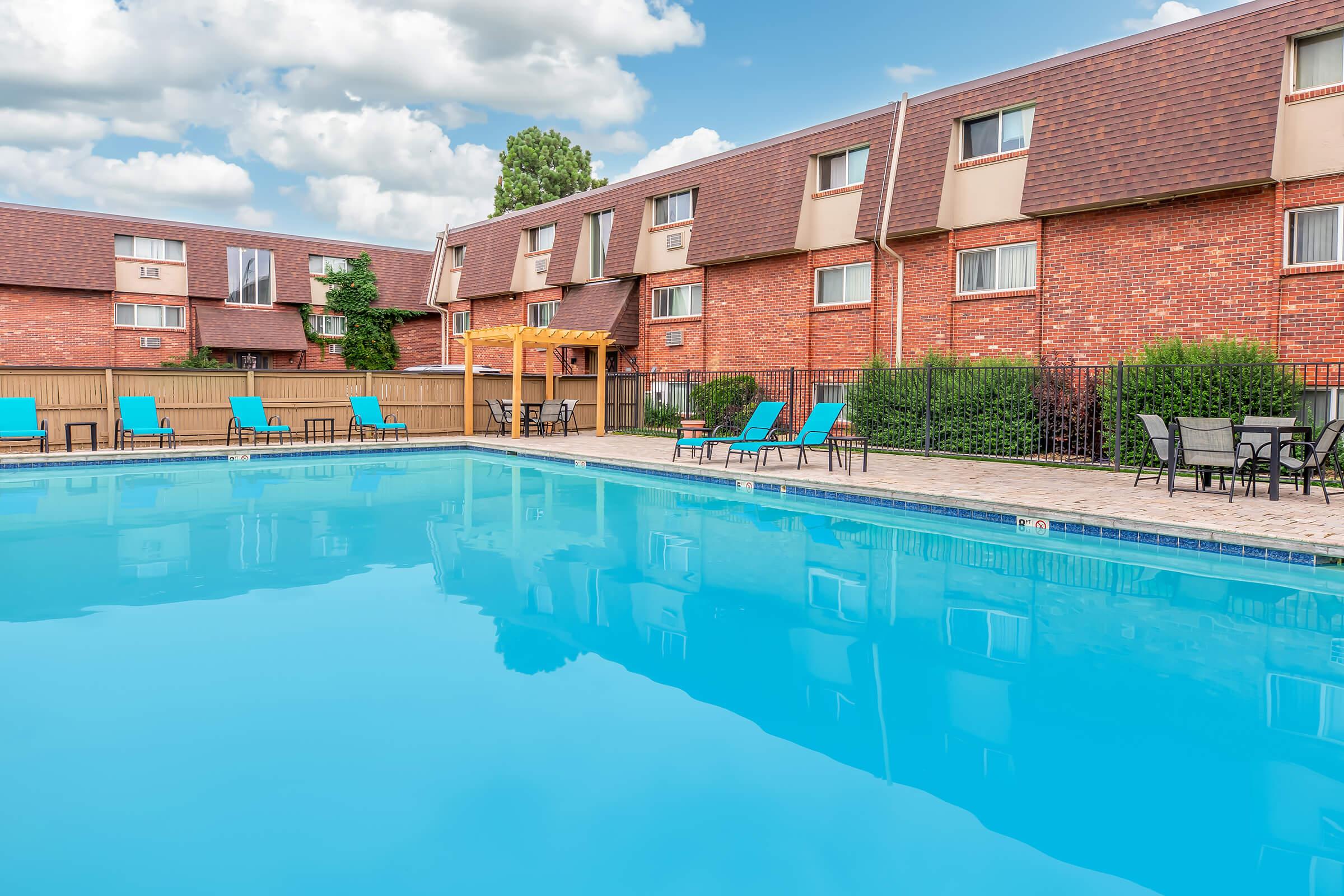A clear blue swimming pool surrounded by lounge chairs and a shaded seating area. The pool reflects the sky and nearby buildings, with well-maintained landscaping and a row of red-brick apartment-style buildings in the background under a partly cloudy sky.