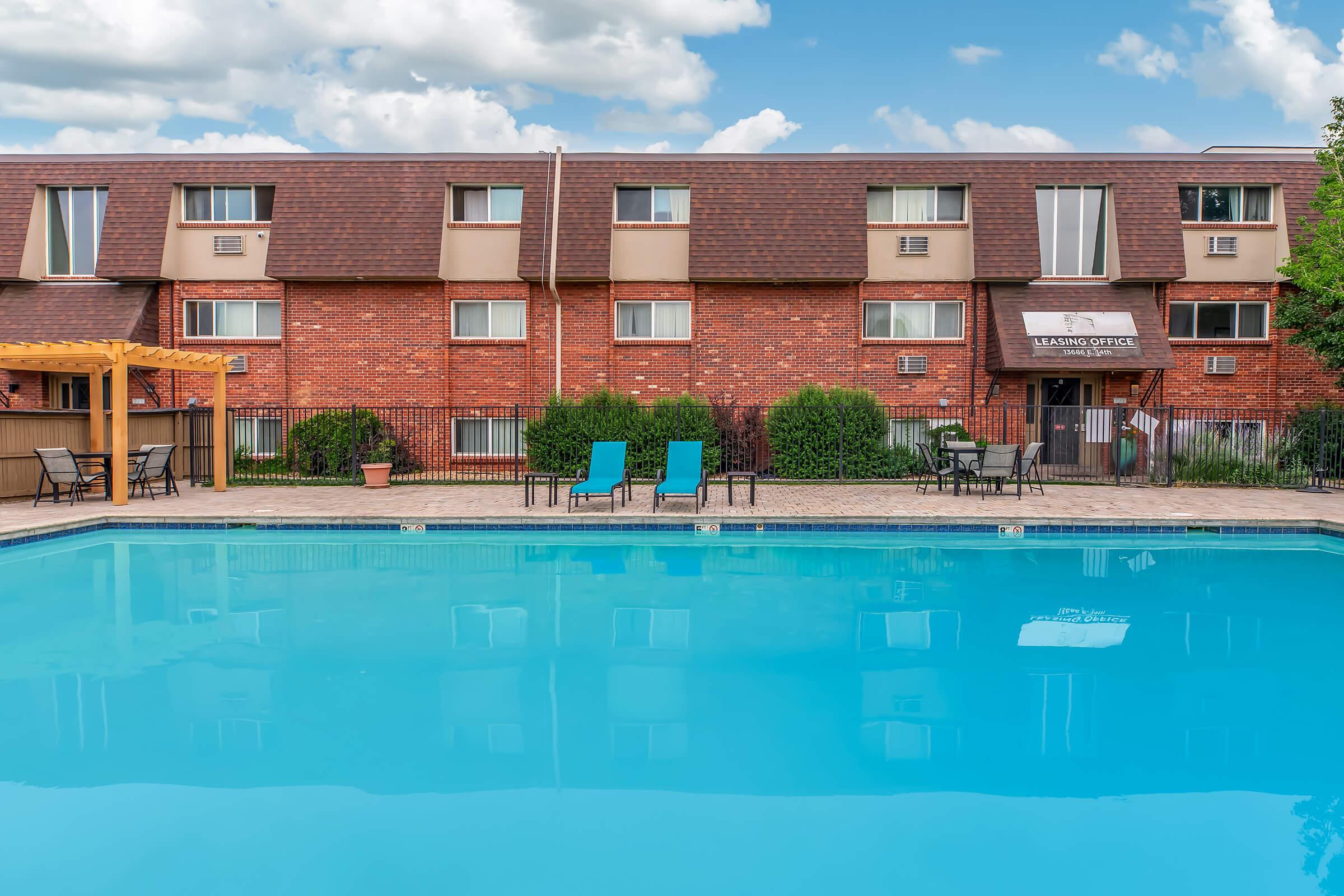A clear blue swimming pool in front of a brick apartment building with a leasing office. Two lounge chairs are placed beside the pool, and there are green shrubs and a pergola in the foreground. The sky is partly cloudy, adding to the inviting atmosphere.