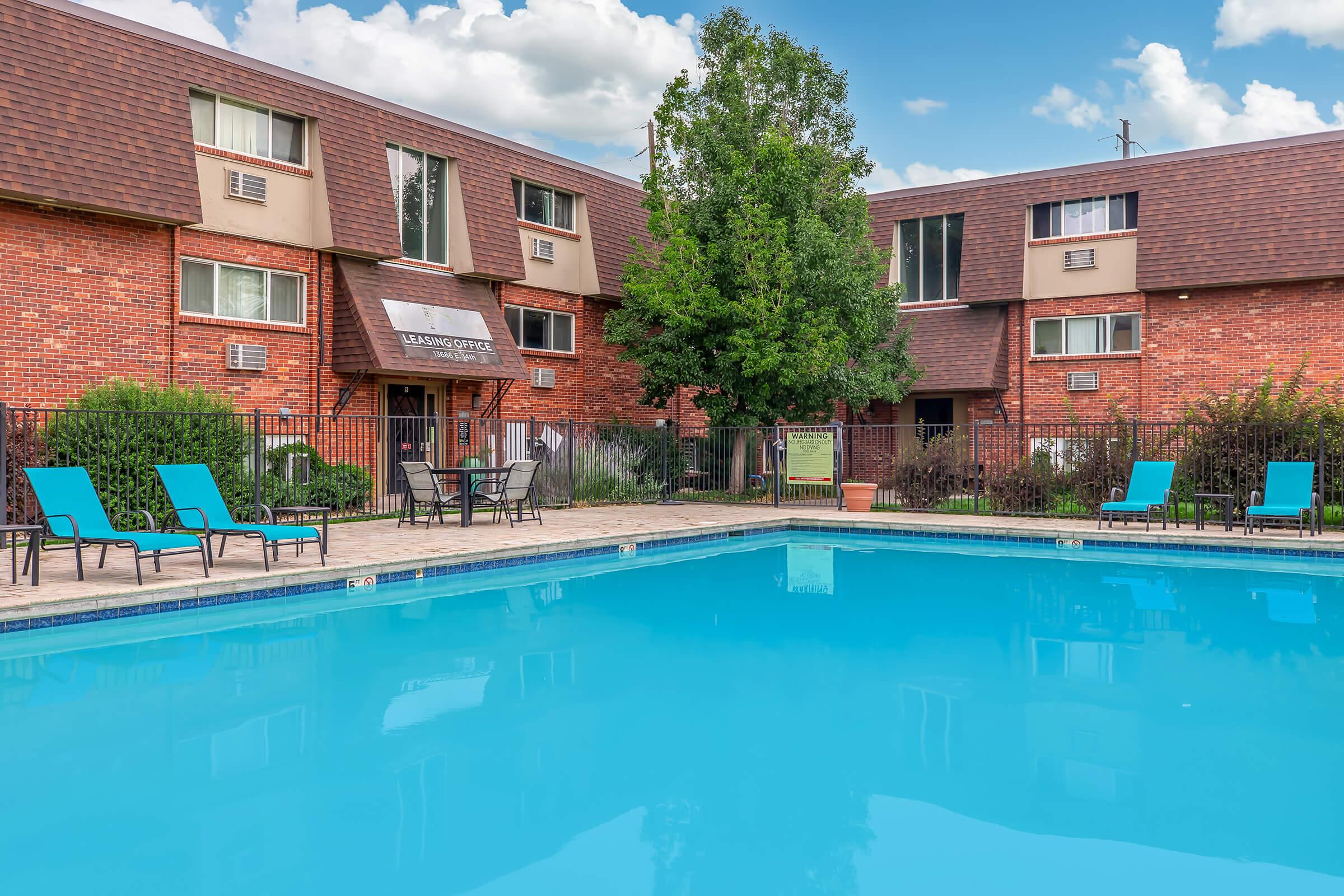 A clear blue swimming pool surrounded by a fenced area, featuring several lounge chairs. In the background, red brick apartment buildings with windows and a sign for the leasing office. Lush greenery adds to the relaxing atmosphere under a partly cloudy sky.