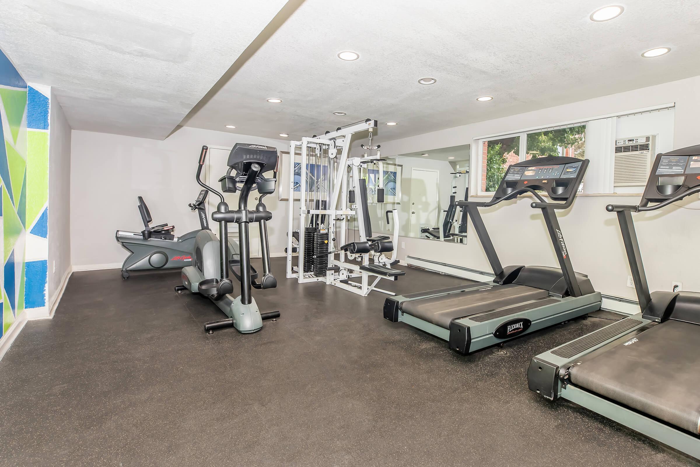 A modern gym interior featuring several exercise machines, including treadmills and an elliptical machine. The space has bright lighting, large windows, and colorful wall accents, creating an inviting atmosphere for workouts.