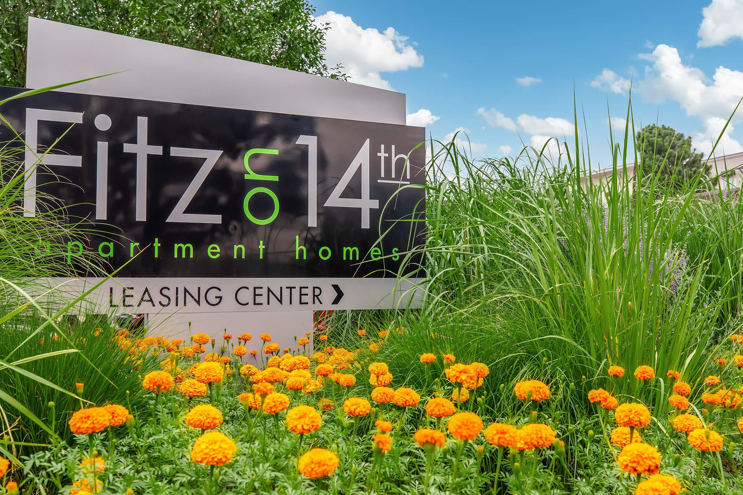 A sign for Fitz on 14th Apartment Homes, prominently displayed in a garden setting with vibrant orange marigold flowers and lush green grass. The sign includes the words "Leasing Center," set against a background of blue sky and fluffy white clouds.
