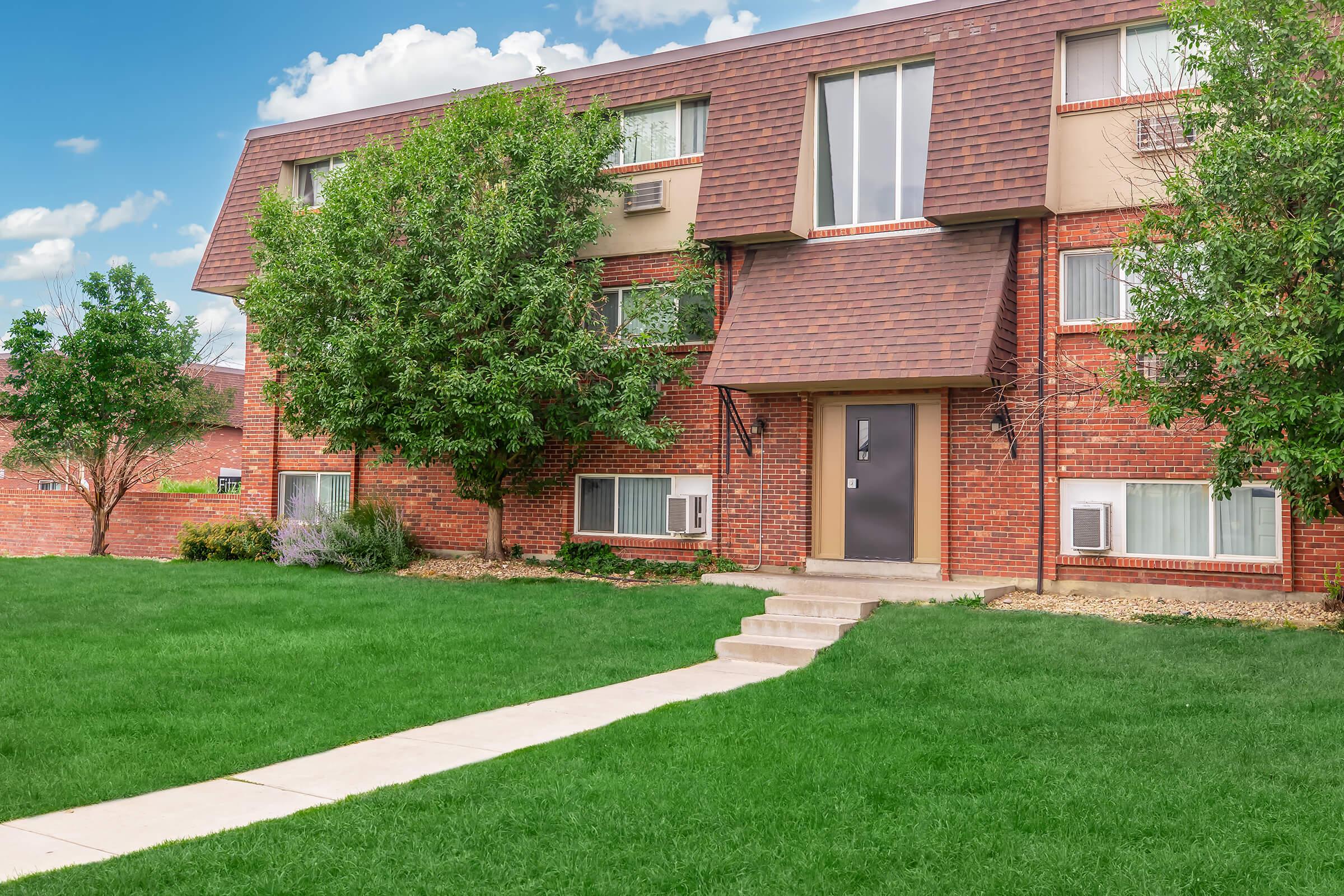 A multi-story brick apartment building with a sloped roof, surrounded by green grass and trees. A concrete pathway leads to the entrance, with two air conditioning units visible on the exterior walls. The sky is partly cloudy, adding a bright, welcoming atmosphere to the scene.