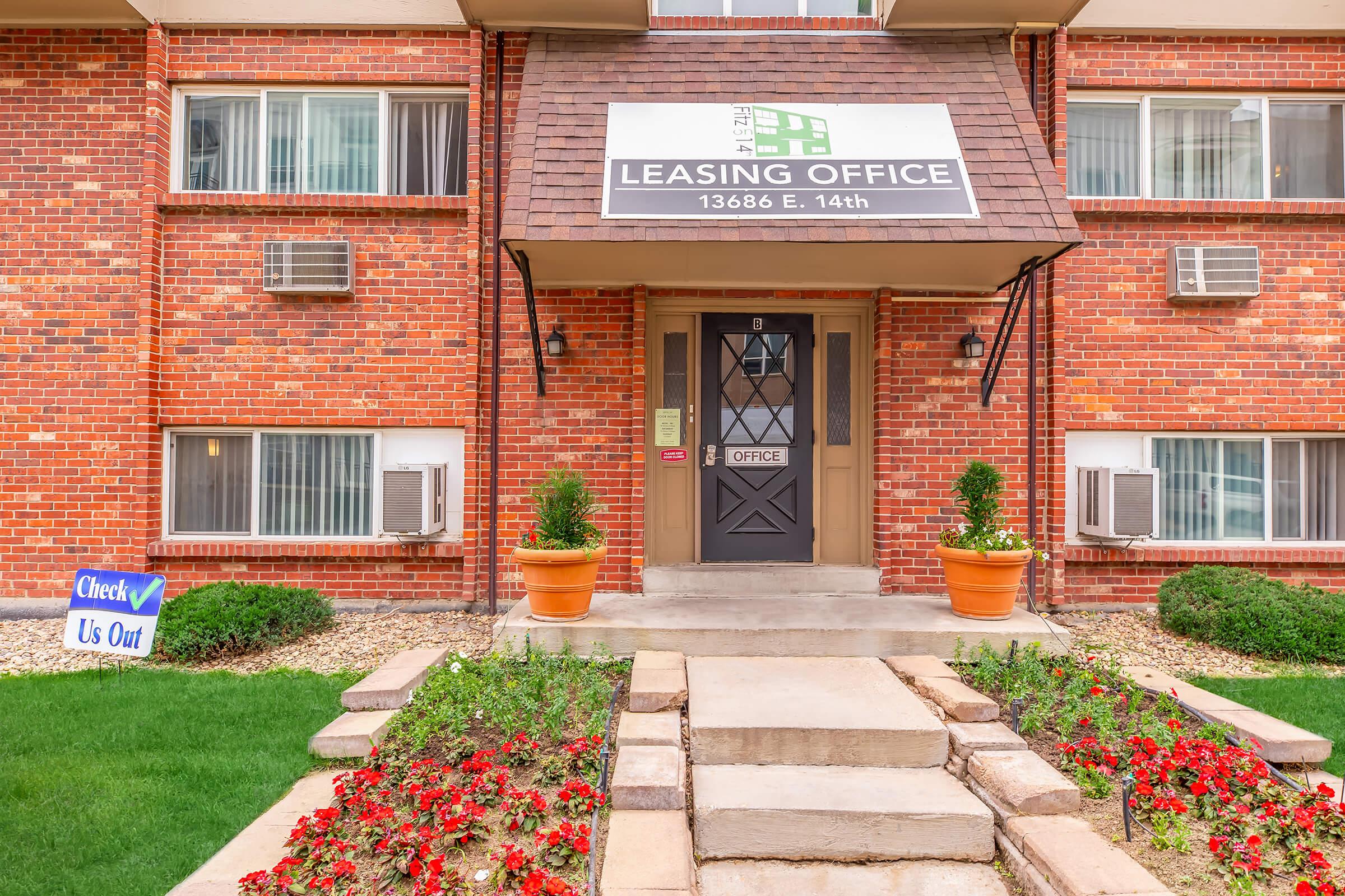Leasing office entrance for an apartment building, featuring a prominent sign that reads "Leasing Office," surrounded by potted plants and a flowerbed. There are air conditioning units on either side of the door, with a welcoming pathway leading to the front entrance. A sign on the lawn invites visitors to "Check Us Out."