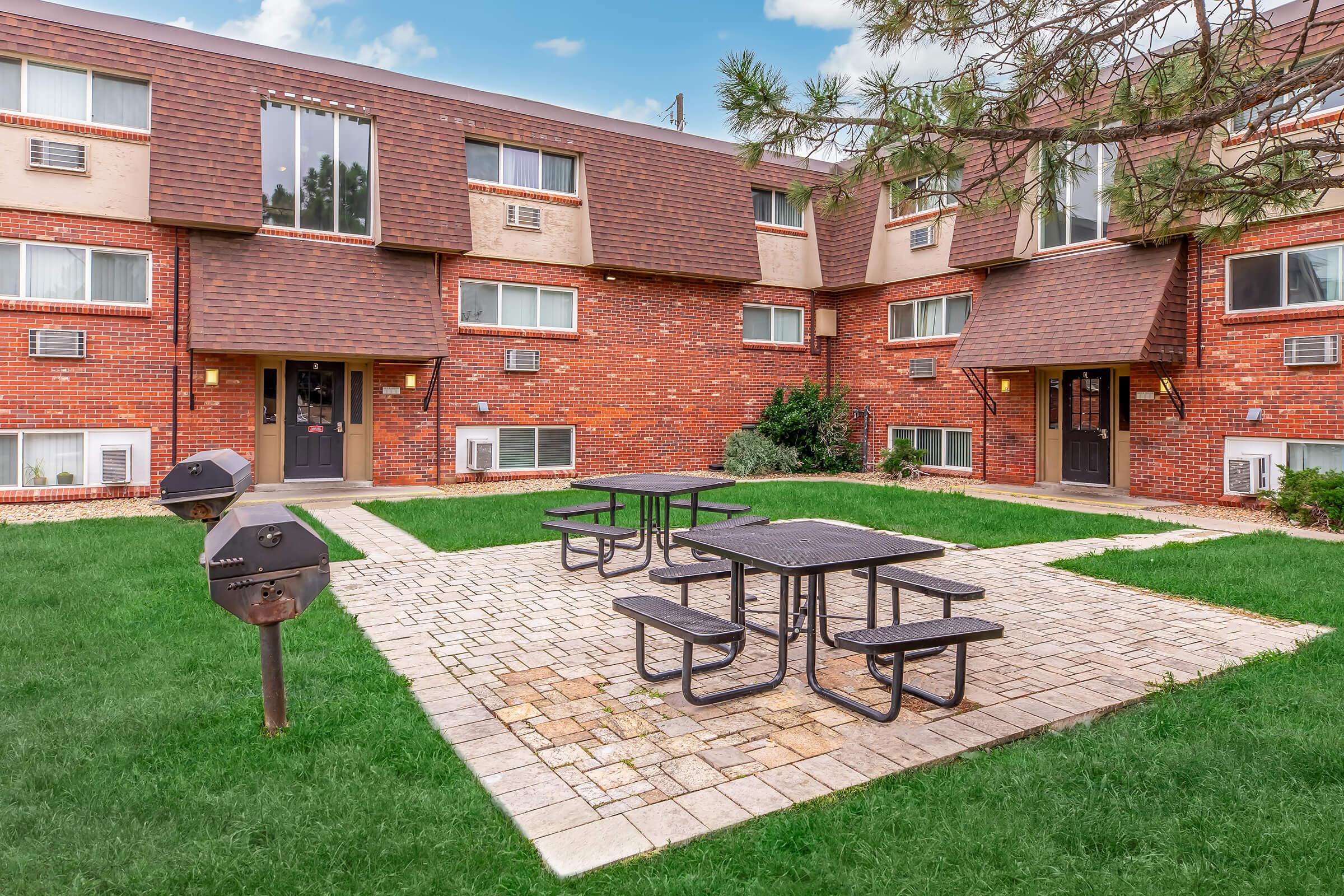 Outdoor common area featuring a paved seating area with black picnic tables and charcoal grills, surrounded by green grass and brick apartment buildings with brown shingled roofs. The scene is bright and inviting, showcasing a space for residents to relax and socialize.