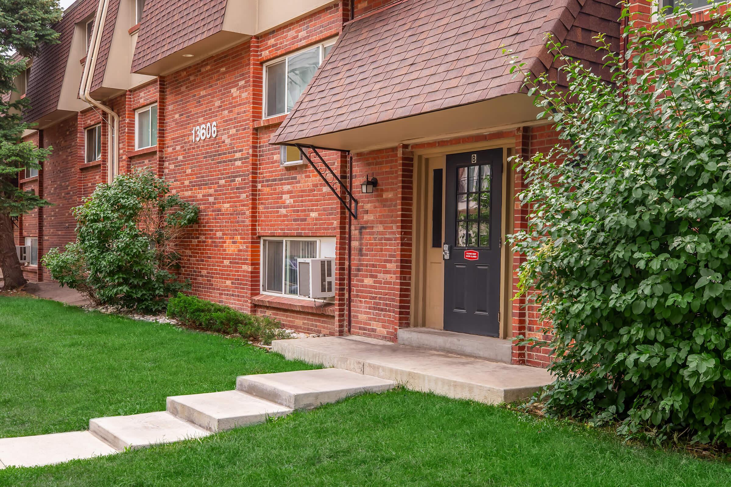 Exterior view of a brick residential building featuring a black front door with a small porch. The entrance is flanked by green shrubs and grass, with concrete steps leading up to the door. Several windows are visible, and the building has a sloped roof with brown shingles.