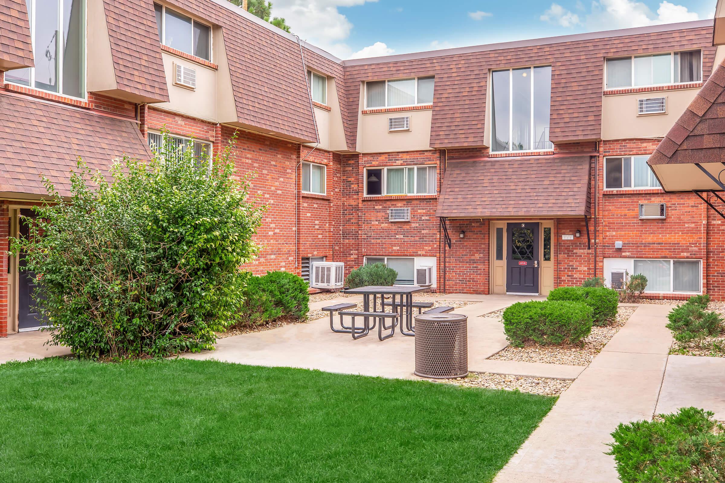 A landscaped courtyard of an apartment complex featuring several brick buildings. There is a patch of green grass, neatly trimmed bushes, a picnic table, and a trash receptacle. The pathway is lined with decorative stones, and air conditioning units are visible in the windows.