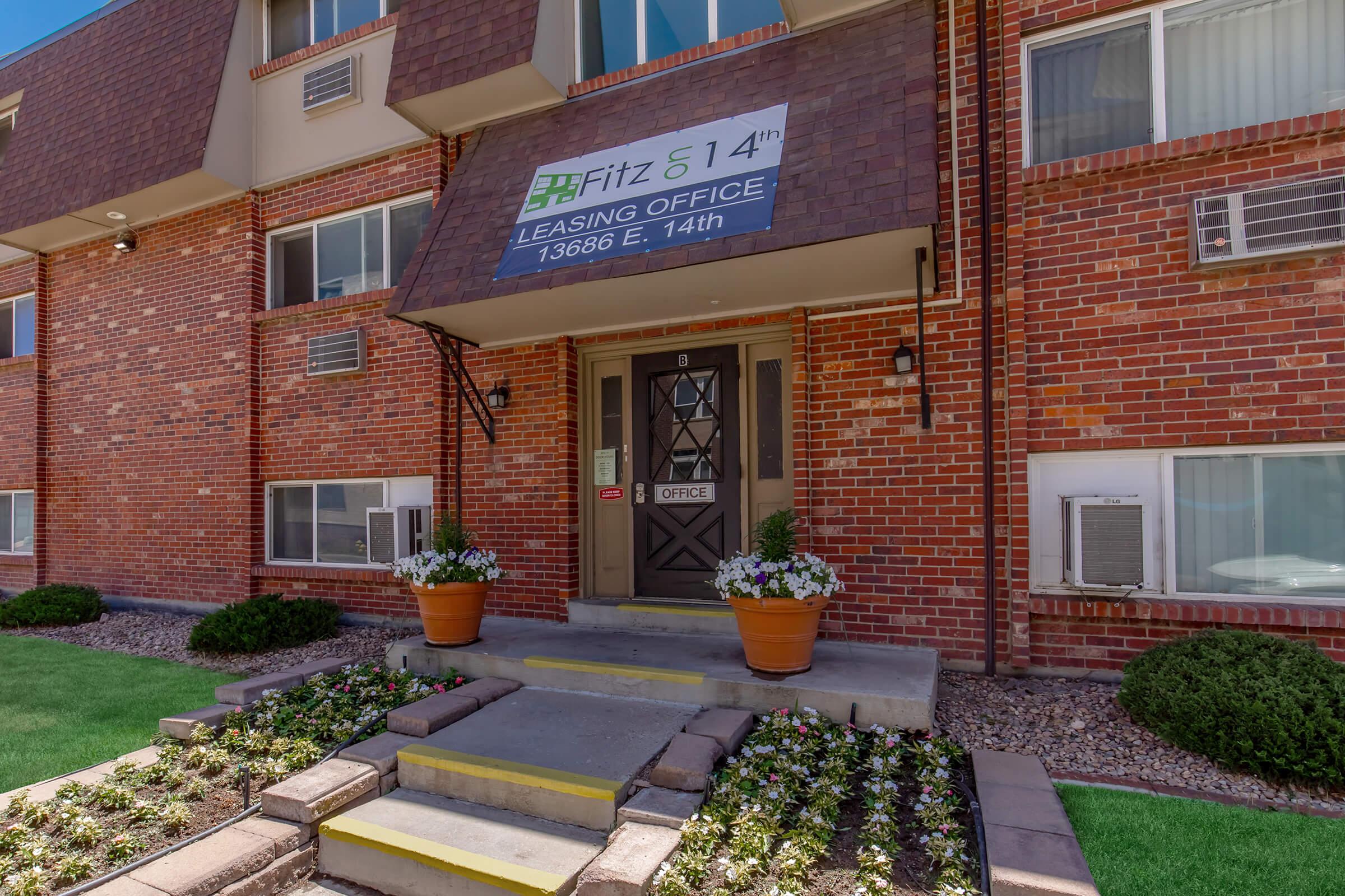 Leasing office of Fitz 514, featuring a brick exterior with large windows. The entrance is marked by a sign reading "Fitz 514 Leasing Office" and has stone steps leading up to a door. Flower pots flank the entrance, and well-maintained flower beds are visible in front of the building.