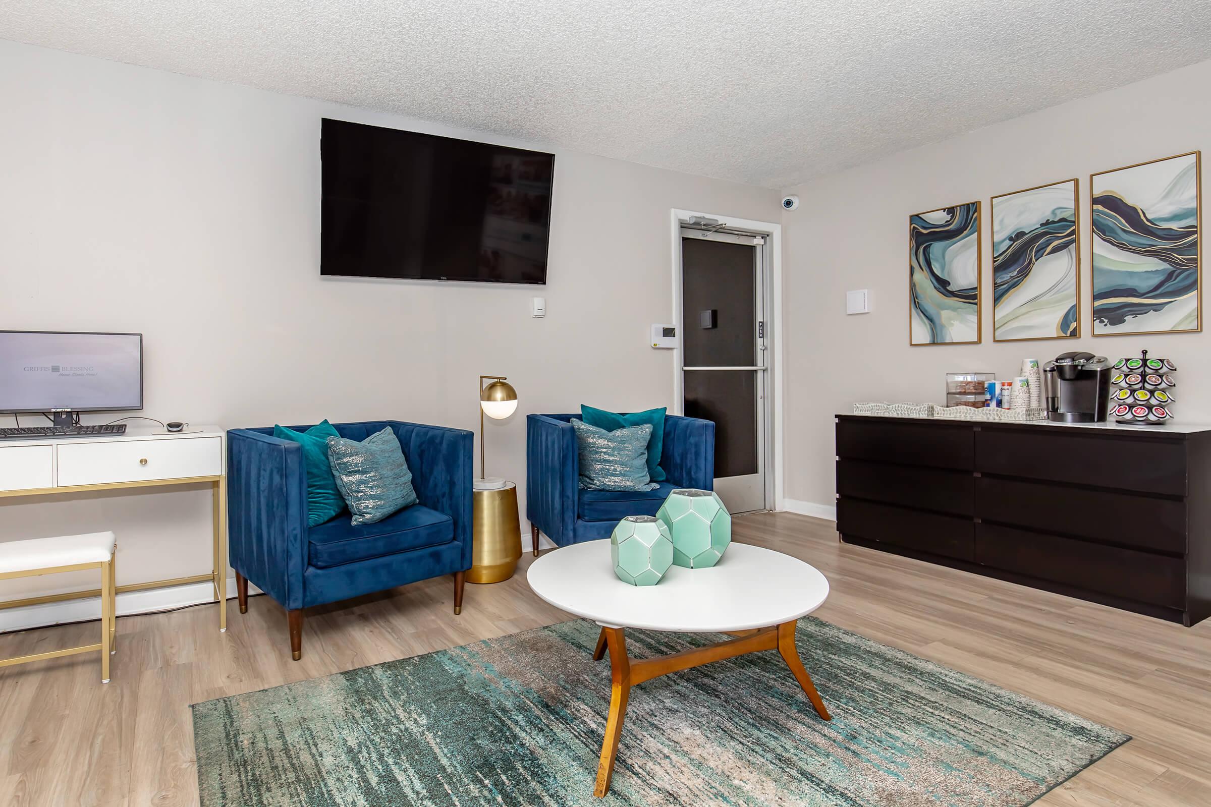 A modern interior of a living room featuring two blue armchairs with decorative pillows, a round coffee table, and a TV mounted on the wall. There's a side table with a lamp and a dark wooden dresser displaying a coffee maker and decor items. A rug adds color to the floor.