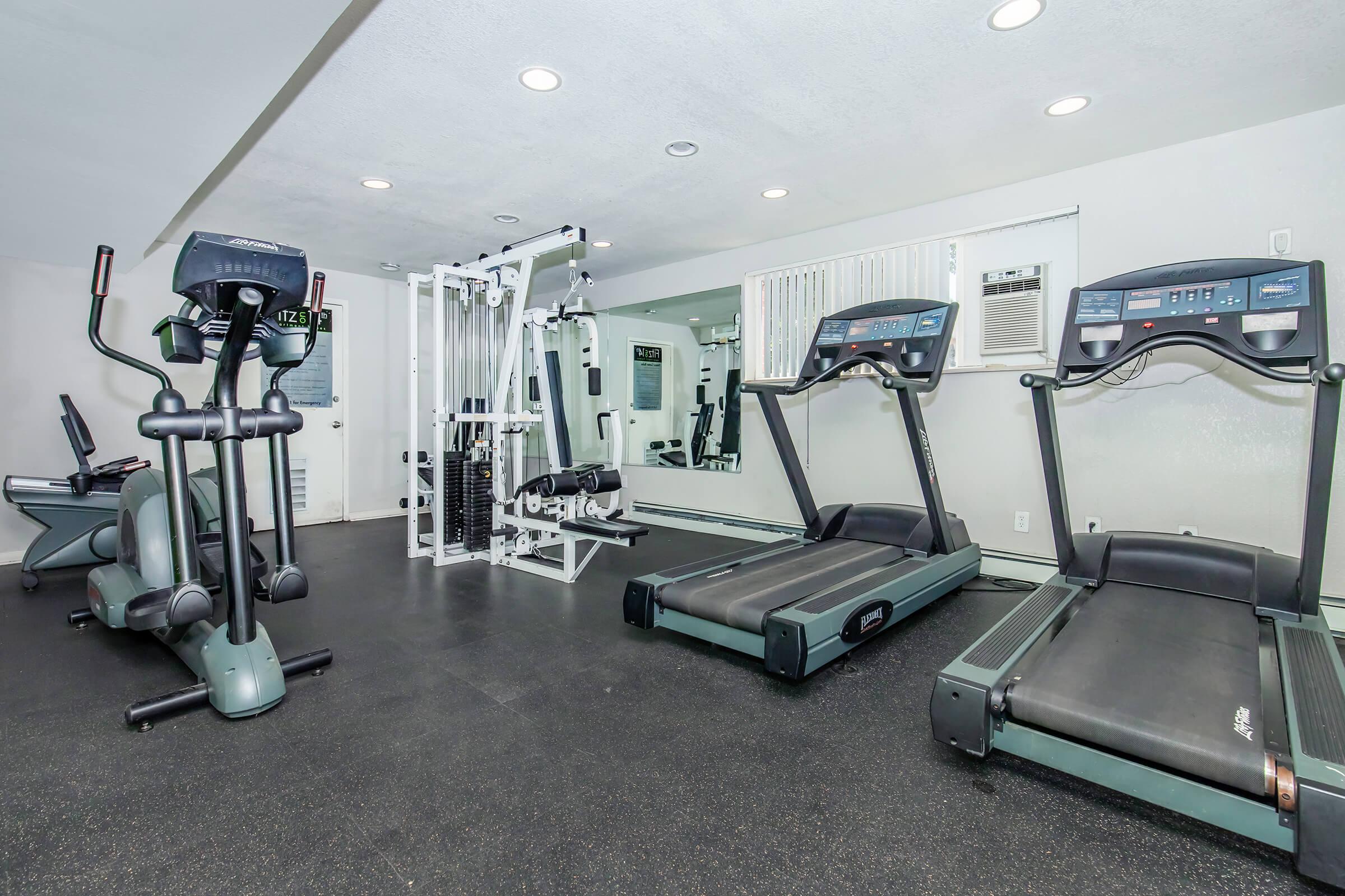 A modern fitness room featuring two treadmills, an elliptical machine, and a multi-station strength training device. The space is well-lit with mirrors on one wall and rubber flooring, creating a clean and inviting exercise environment.
