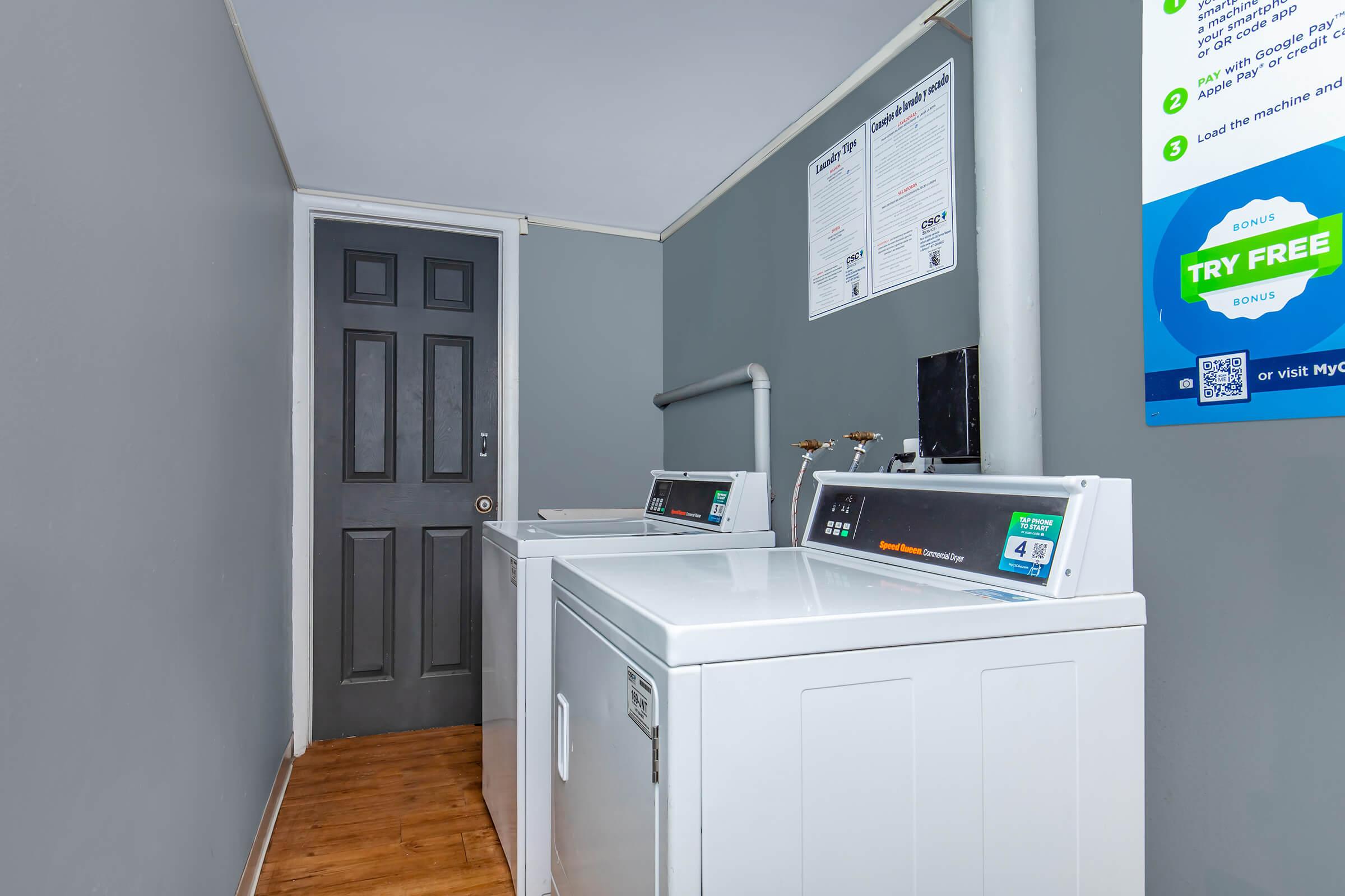 A laundry room featuring two white washing machines against a gray wall. There is a dark door at the end of the room and a bulletin board with laundry instructions and promotions. The wood floor adds warmth to the space.