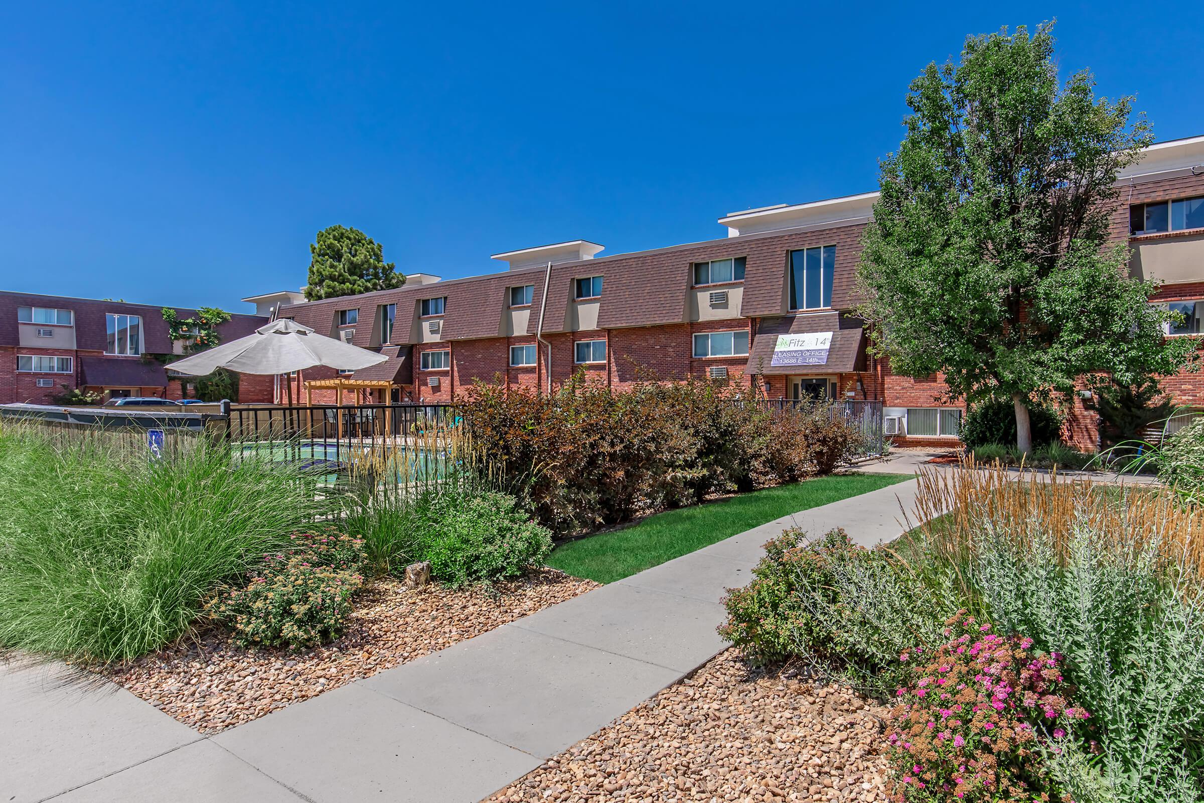 A well-maintained apartment complex with a landscaped pathway, featuring grass and flowering plants. In the foreground, a swimming pool area is visible with an umbrella, and the building has multiple air conditioning units and large windows under a bright blue sky.