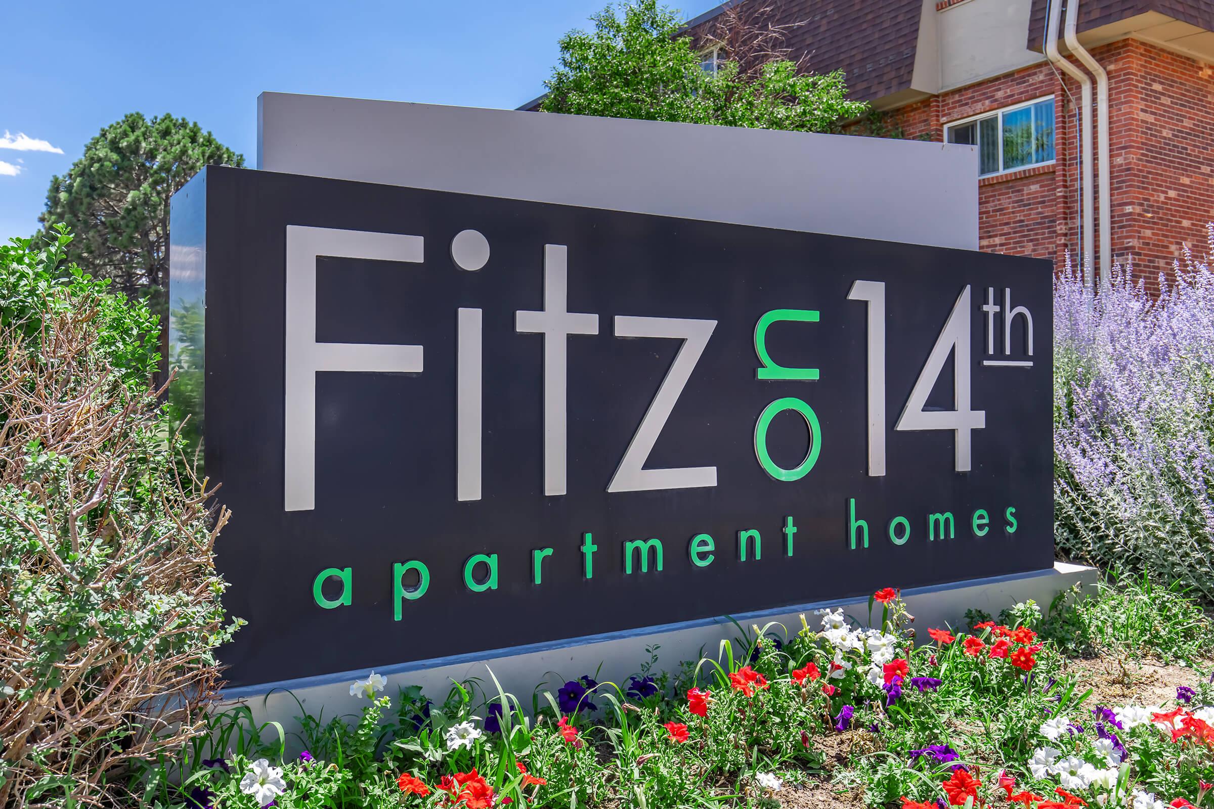 Sign for "Fitz on 14th apartment homes" with vibrant flowers and landscaping around it. The design features modern typography in black and silver against a backdrop of greenery and colorful blooms under a clear blue sky.
