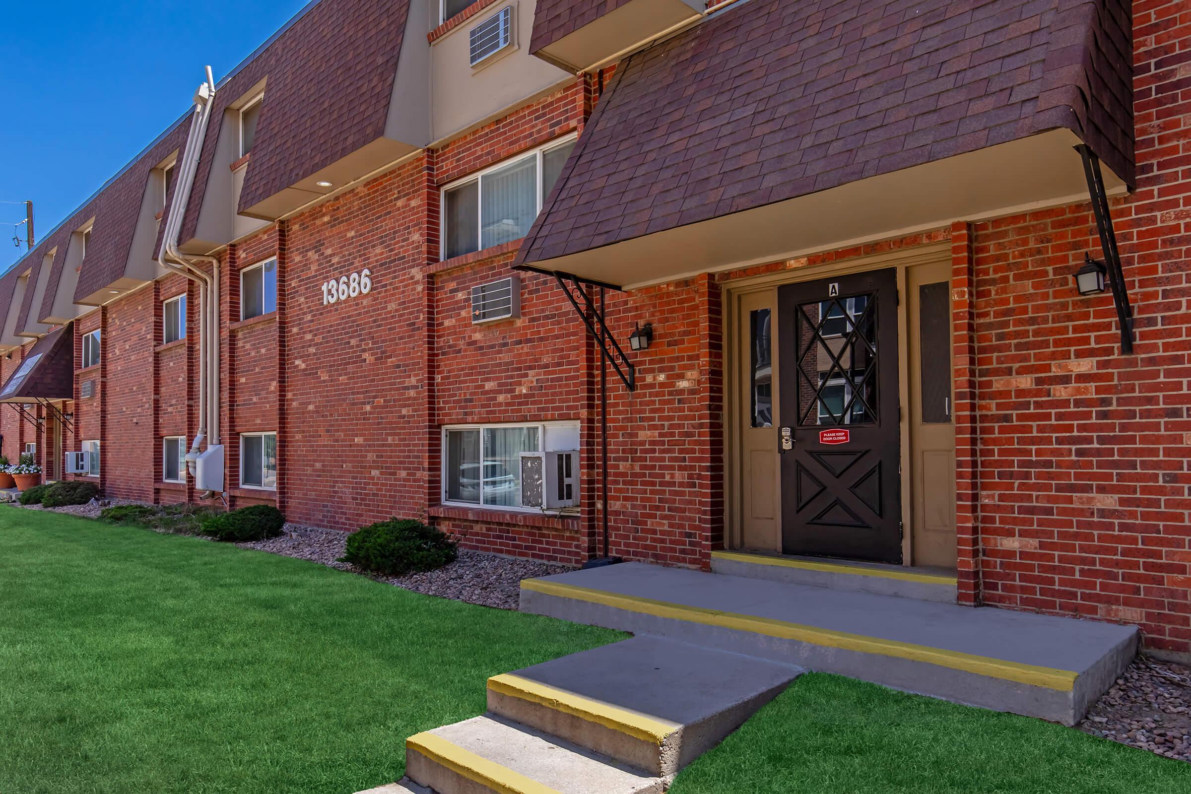Exterior view of a multi-unit brick apartment building featuring a prominent entrance with a brown door, steps leading up to the door, and well-maintained green grass and landscaping. The building has multiple windows and air conditioning units on the exterior.