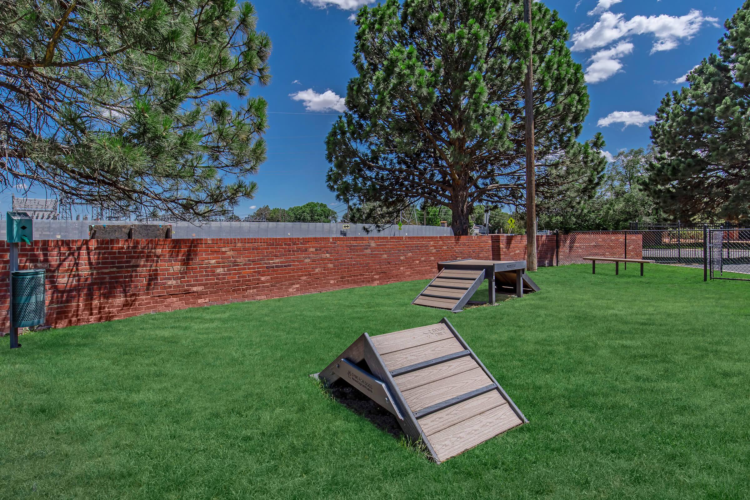 A sunny outdoor area featuring a grassy field with two wooden ramps and a bench. In the background, there are tall trees and a red brick wall, with a clear blue sky and scattered clouds overhead. The space is designed for activities, possibly for pets or recreational use.