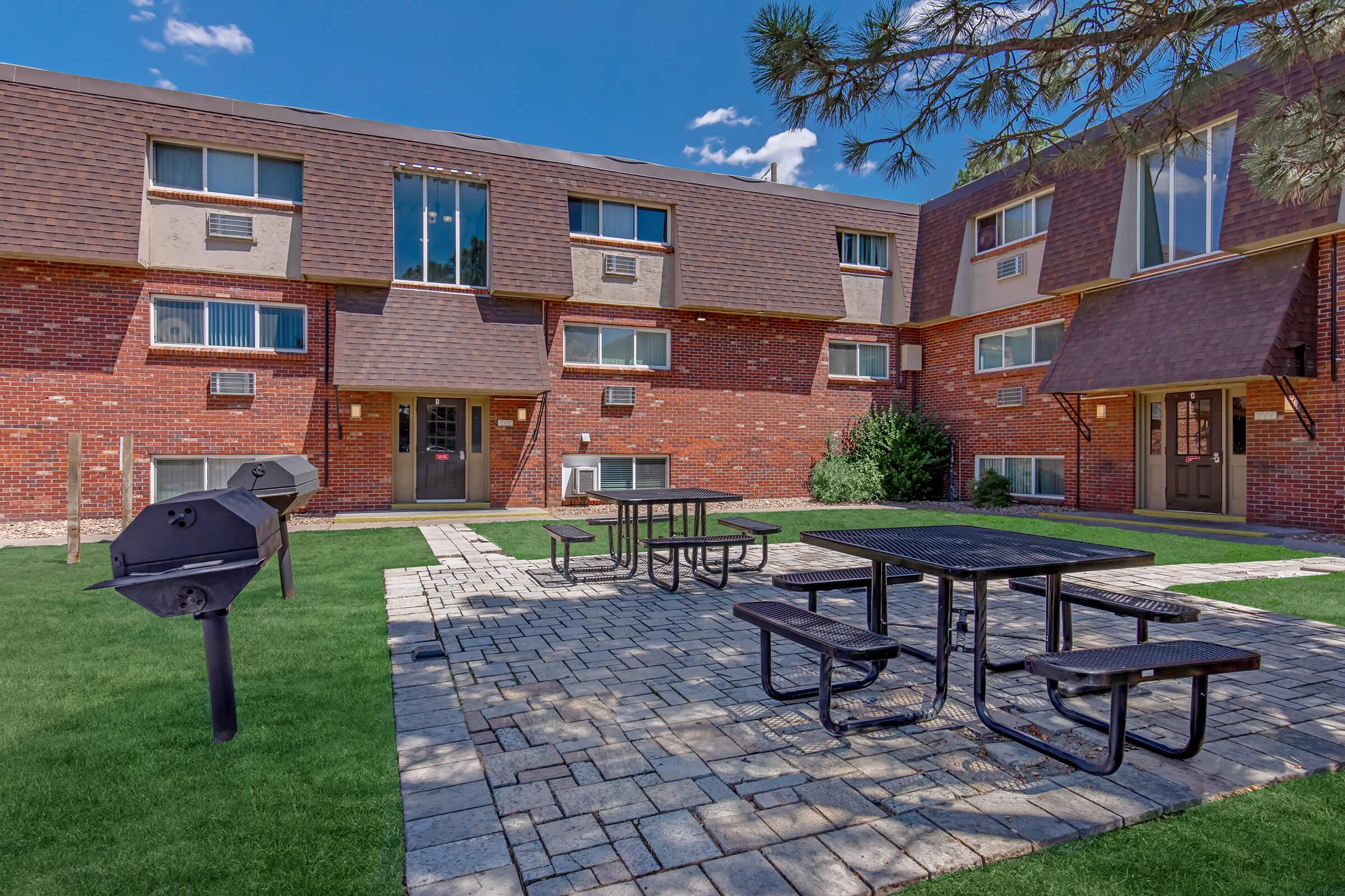 Courtyard area with a brick building in the background featuring multiple windows and air conditioning units. Equipped with several black picnic tables on a paved stone surface, surrounded by green grass. A charcoal grill is positioned on the left side of the image under a tree. Clear blue sky above.