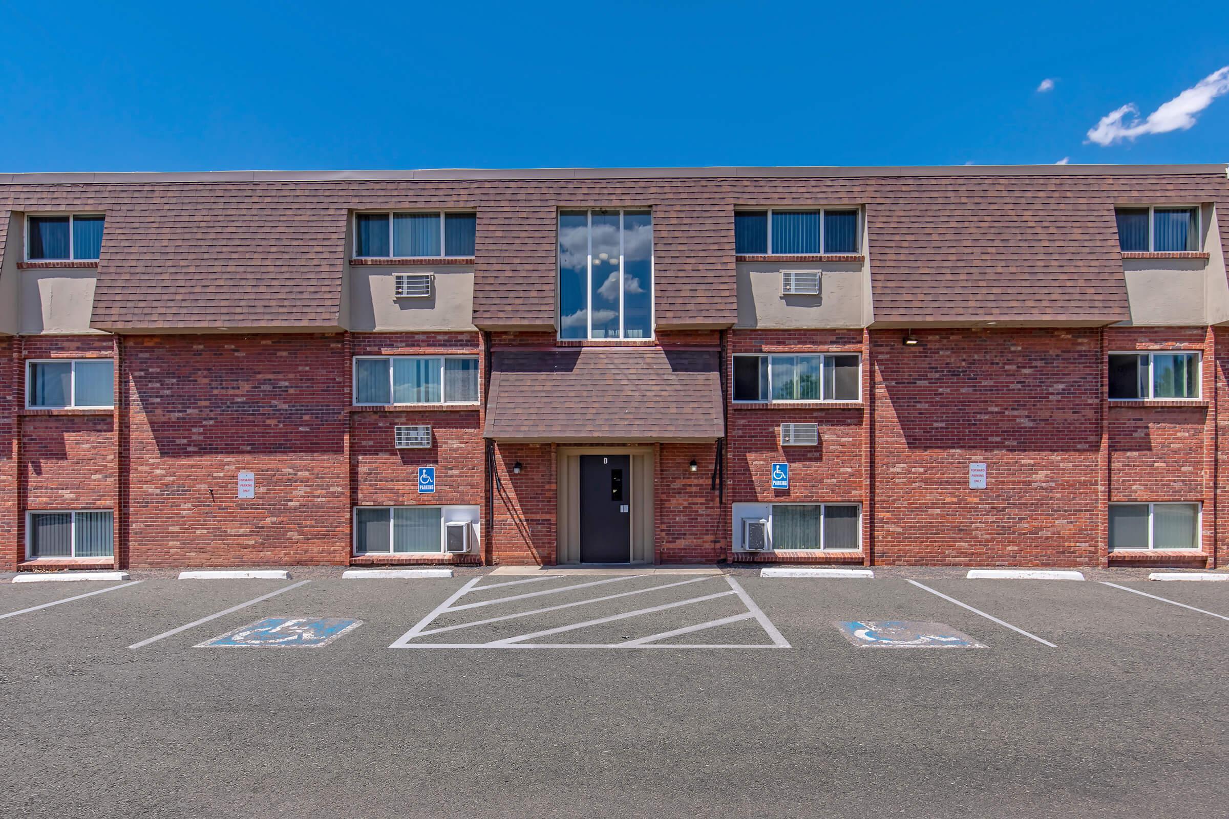 A two-story brick apartment building with a flat roof and large windows. The entrance features a central door and multiple air conditioning units mounted outside. In the foreground, there are designated parking spaces, including accessible spots, under a clear blue sky.