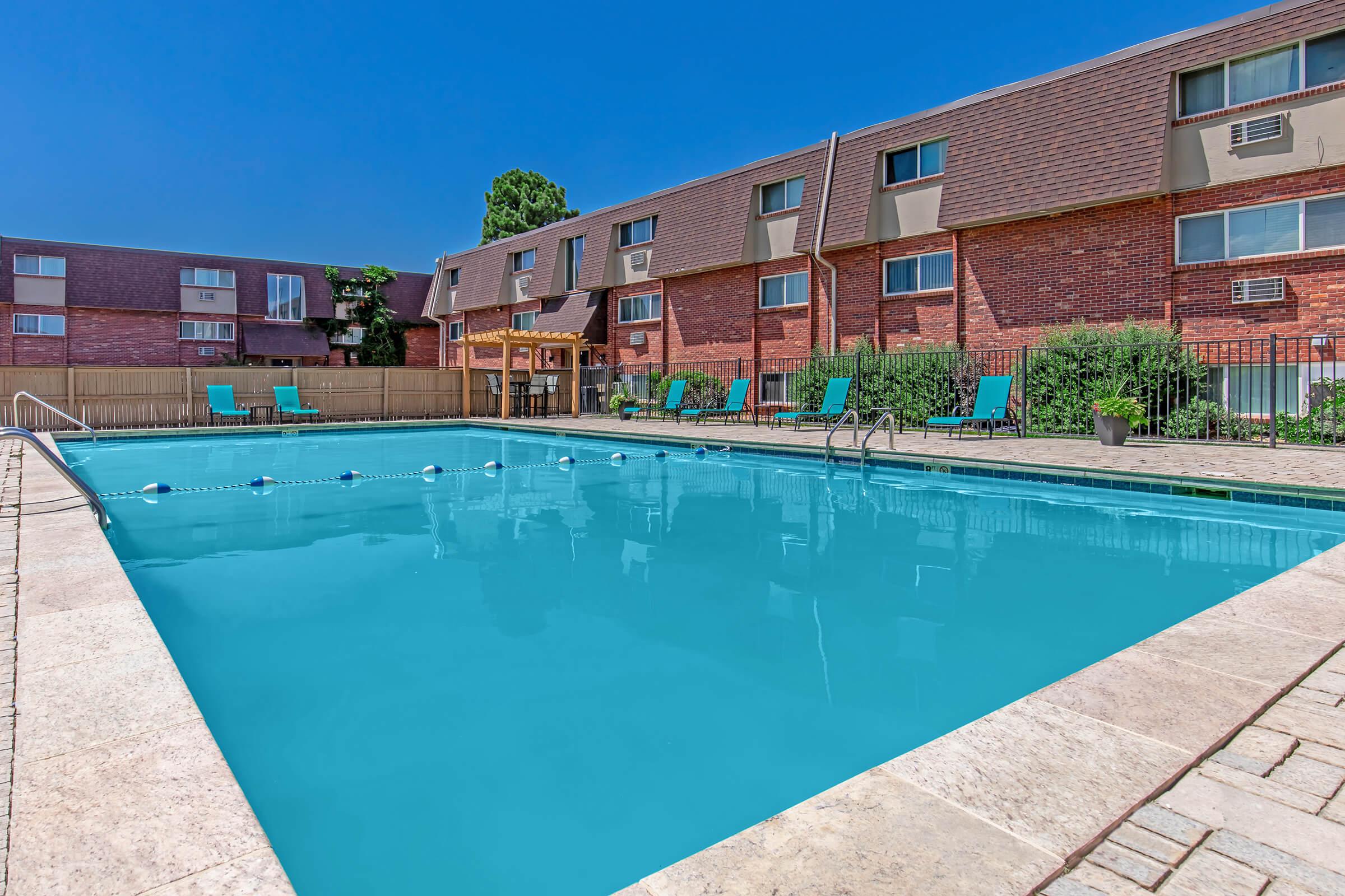 A clear blue swimming pool surrounded by lounge chairs, with a wooden pergola nearby. The pool area is bordered by a fence and there are rows of red-brick apartment buildings in the background under a clear blue sky.