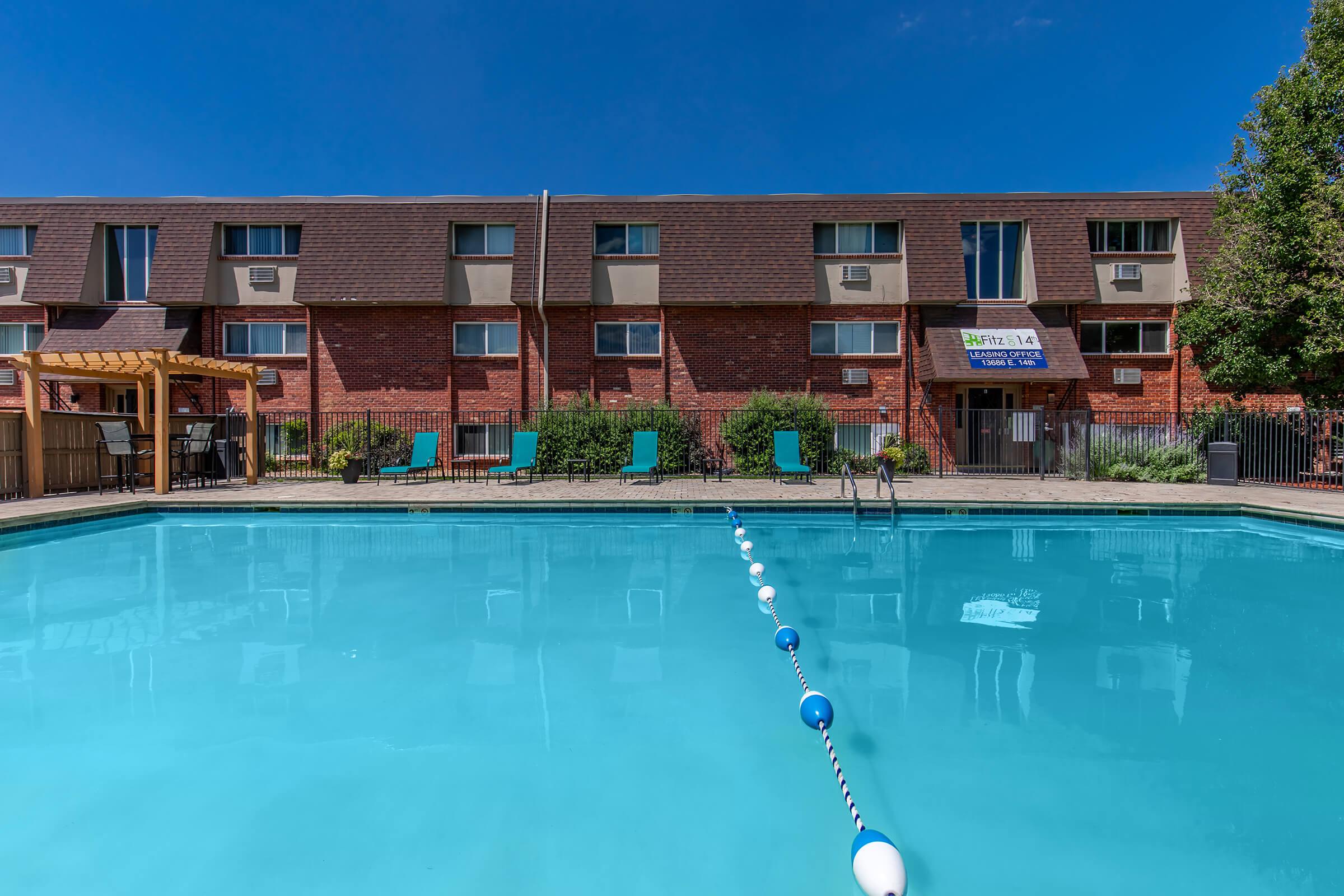 A clear blue swimming pool in the foreground, featuring a lane marker. In the background, a two-story red-brick building with multiple windows, some air conditioning units, and green lounge chairs arranged around the pool area. The sky is bright blue with a few clouds.
