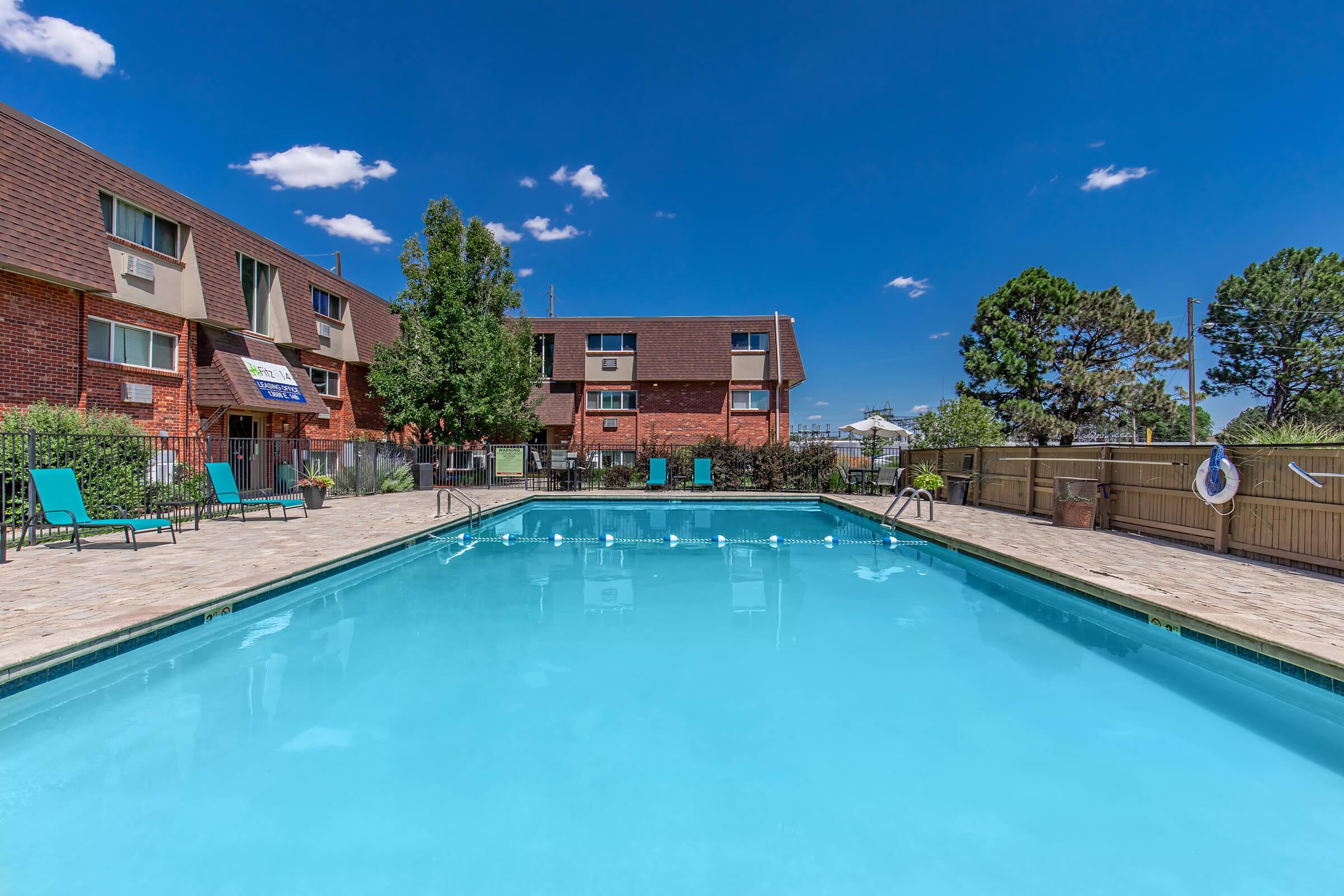 A clear swimming pool surrounded by lounge chairs on a patio. In the background, there are brick buildings with trees and a blue sky dotted with clouds. The pool area is well-maintained and inviting, ideal for relaxation or recreational activities.