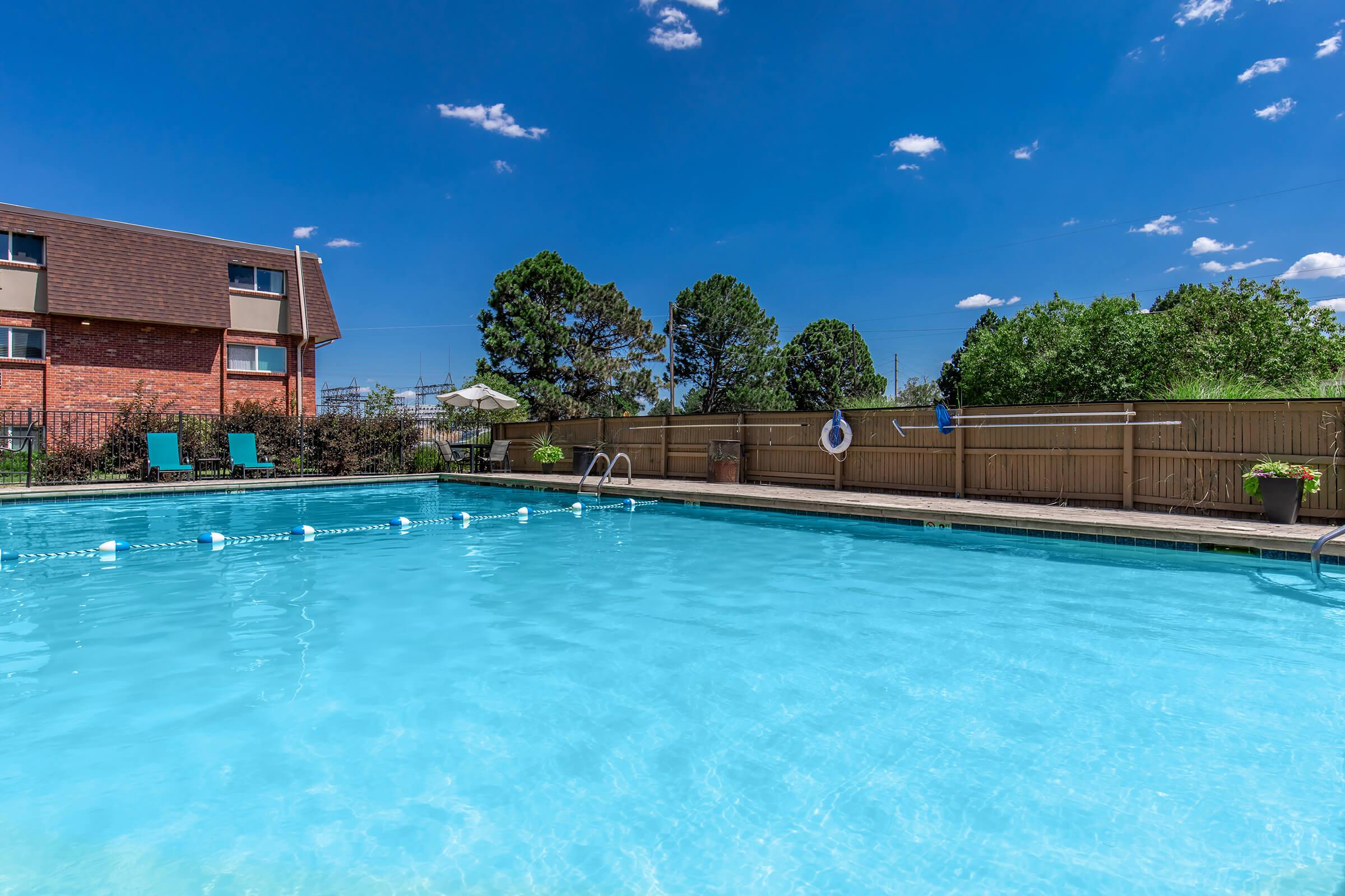 A clear blue swimming pool surrounded by a wooden fence, with a few green trees and blue skies overhead. There are lounge chairs on the poolside, and visible pool accessories like a life buoy and potted plants. The scene conveys a relaxing outdoor atmosphere.