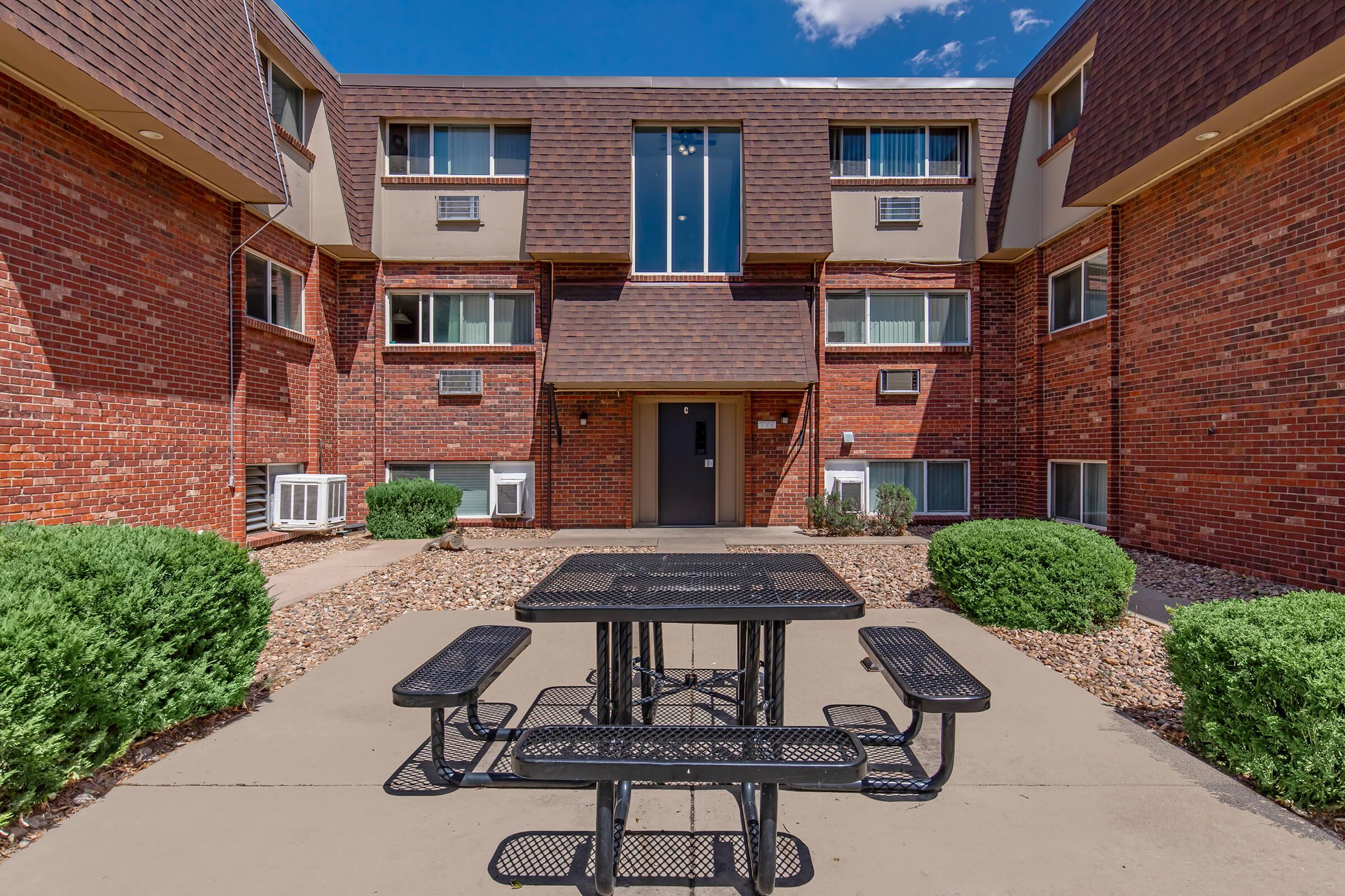 Outdoor area featuring a black metal picnic table with benches, surrounded by well-maintained shrubs and gravel. The background shows a brick building with windows and air conditioning units, under a clear blue sky. The setting is inviting and suitable for social gatherings.