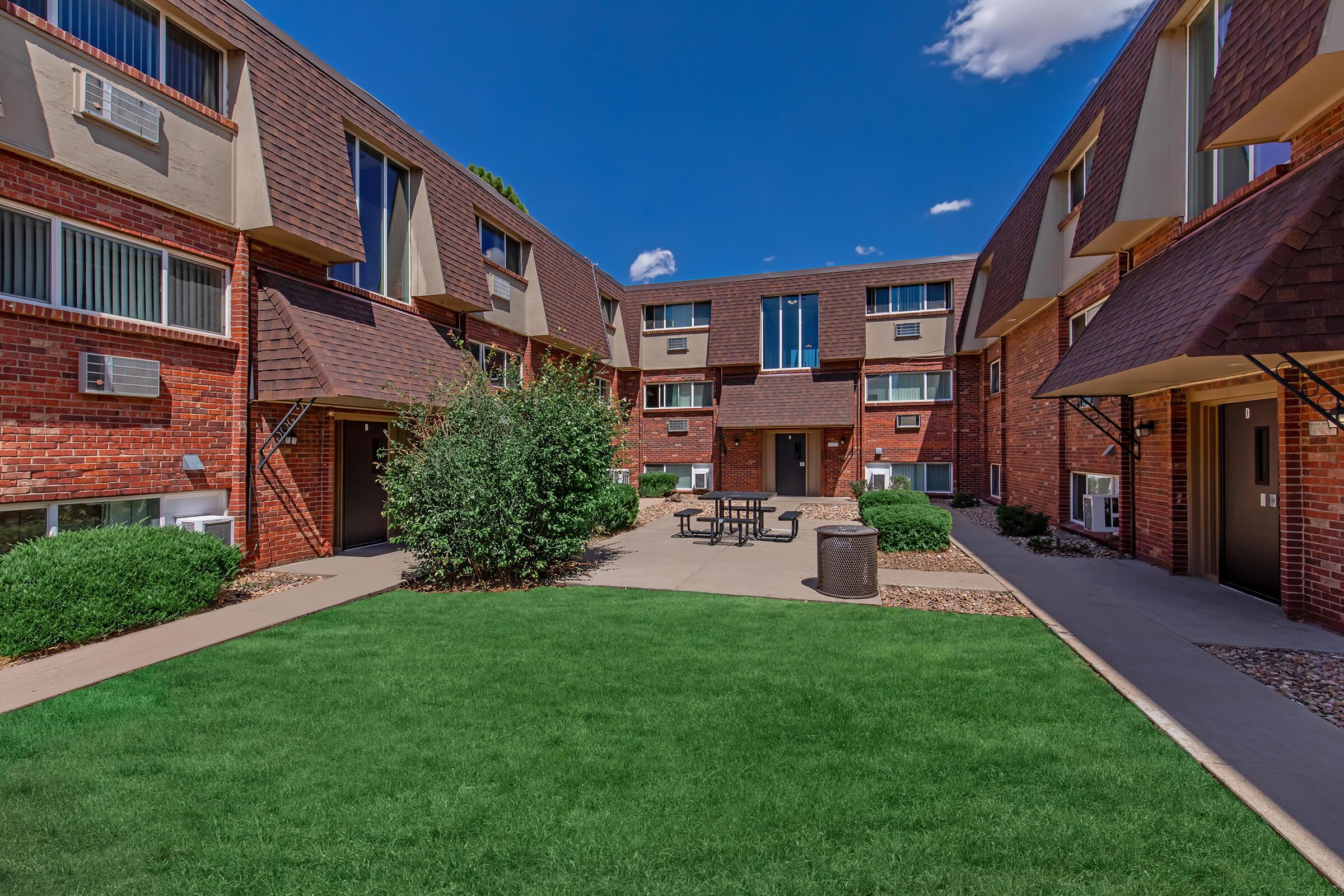 A well-maintained courtyard surrounded by brick apartment buildings. The area features a neatly manicured lawn, a set of outdoor tables and chairs, and several trees and shrubs. The sky is clear with a few scattered clouds, creating a pleasant atmosphere in a residential setting.