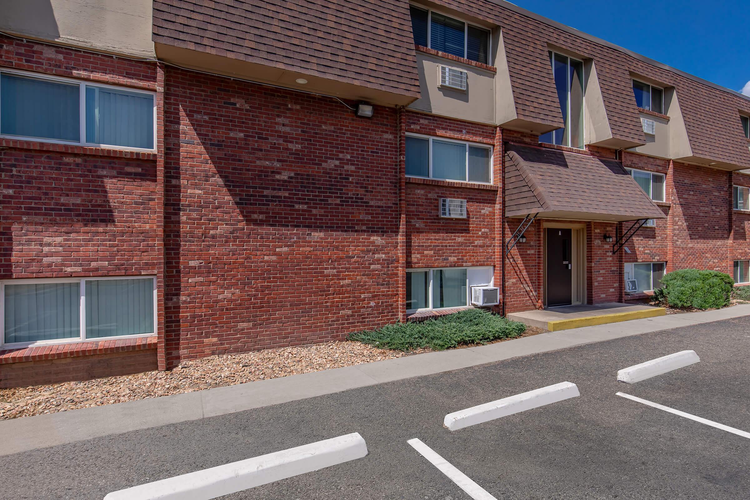 Exterior view of a two-story brick building with large windows and a sloped roof. The entrance is at the center, flanked by air conditioning units. In the foreground, there are several white parking spaces on a paved surface, with small bushes and gravel landscaping. Clear blue sky overhead.
