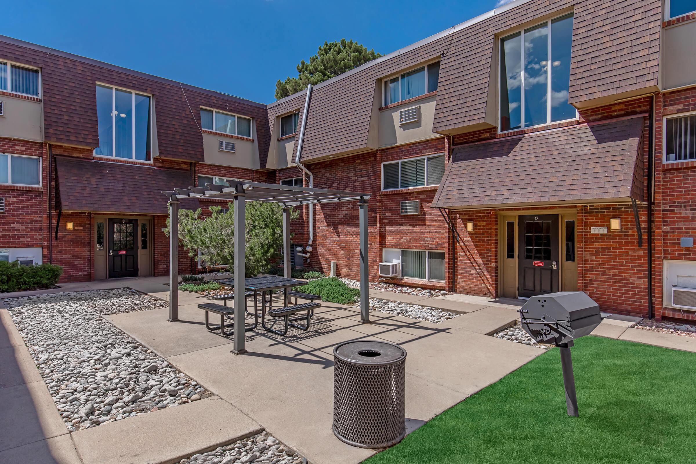 A courtyard area featuring a picnic table under a metal pergola, surrounded by green grass and decorative rocks. There is a grill nearby and a trash can. Two brick apartment buildings with large windows are visible in the background, under a clear blue sky.