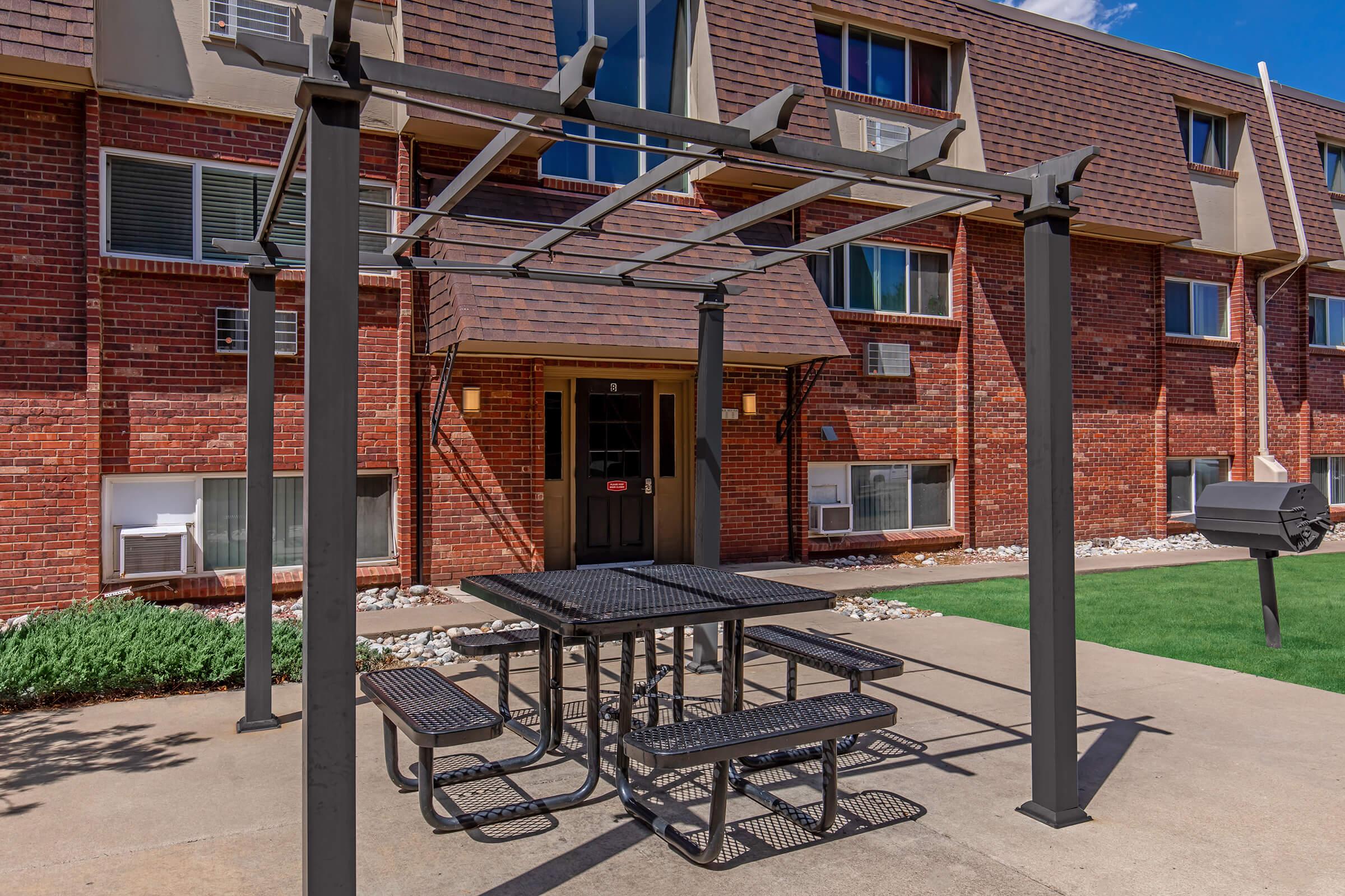 A metal picnic table with attached benches under a pergola, situated in front of a brick apartment building. The building features multiple windows and air conditioning units, with a well-maintained lawn and pathway leading to the entrance.