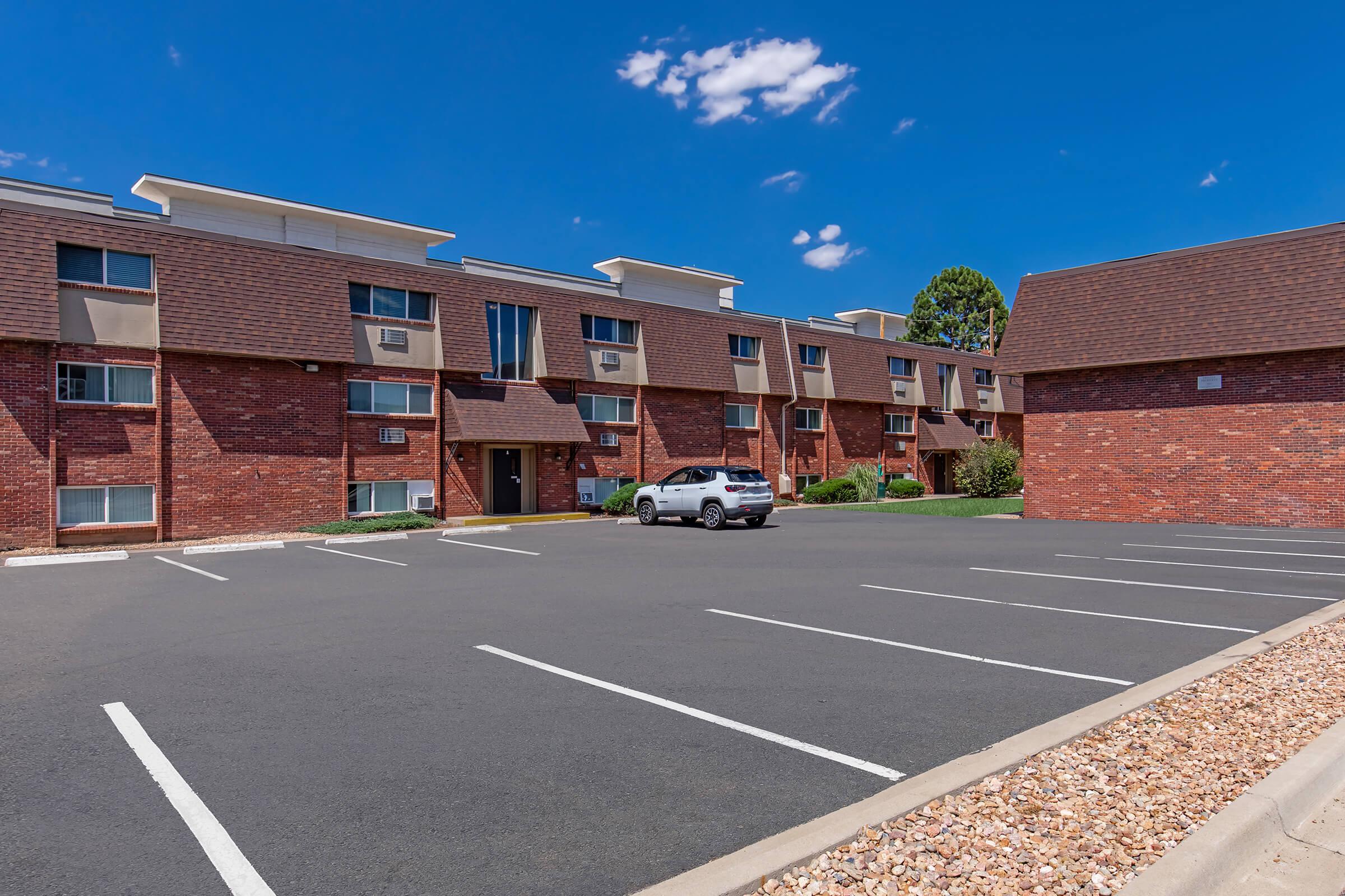 A modern brick apartment building featuring multiple units. The exterior showcases a mix of brick and siding, with several air conditioning units visible. A clear blue sky with a few clouds is above. The parking lot in front has marked spaces, and a white SUV is parked nearby, surrounded by well-maintained greenery.
