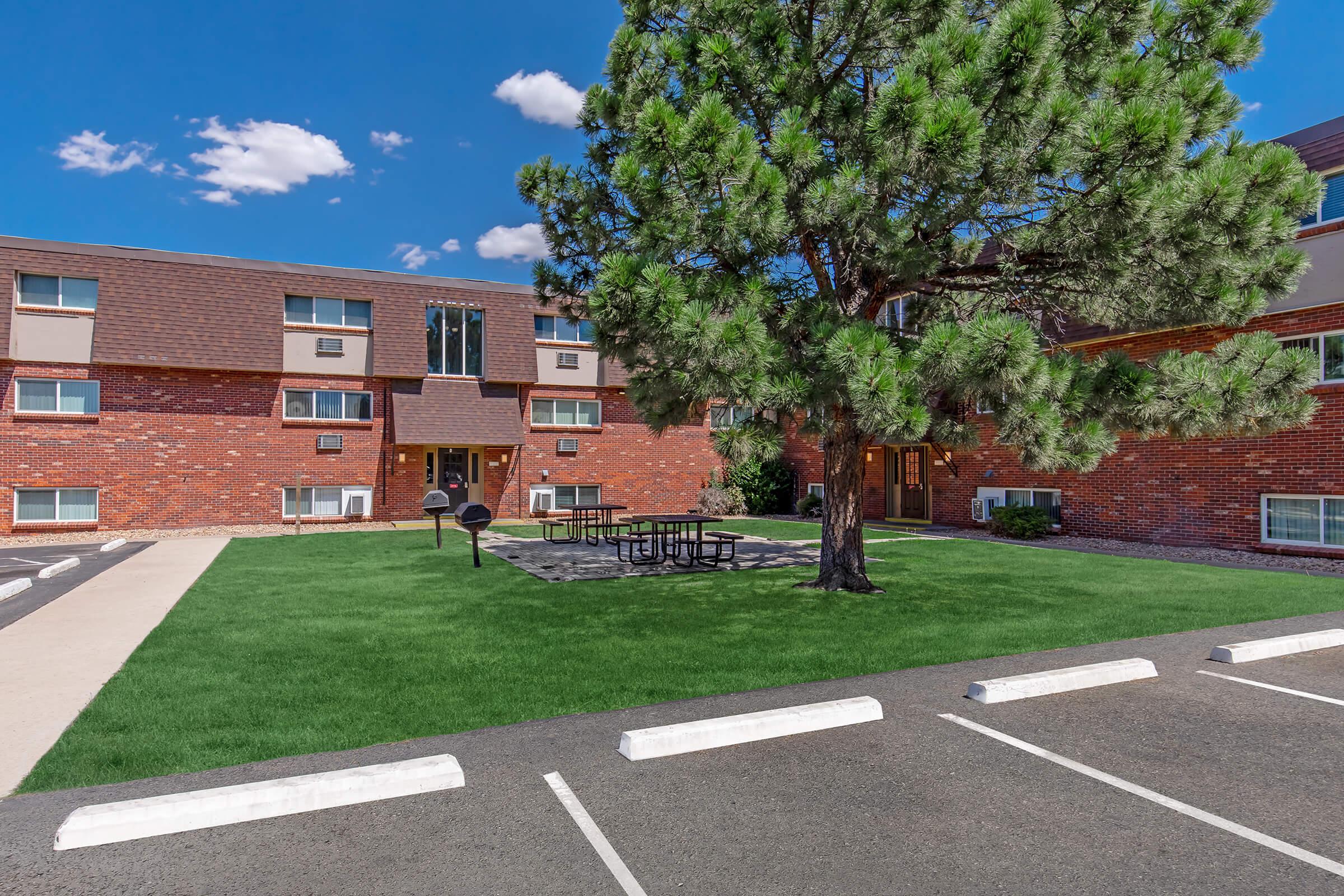 A well-maintained residential building surrounded by green grass and a large tree. There are outdoor picnic tables and parking spaces visible in the foreground. The sky is clear with a few clouds, and the brick facade of the building adds to the welcoming atmosphere.